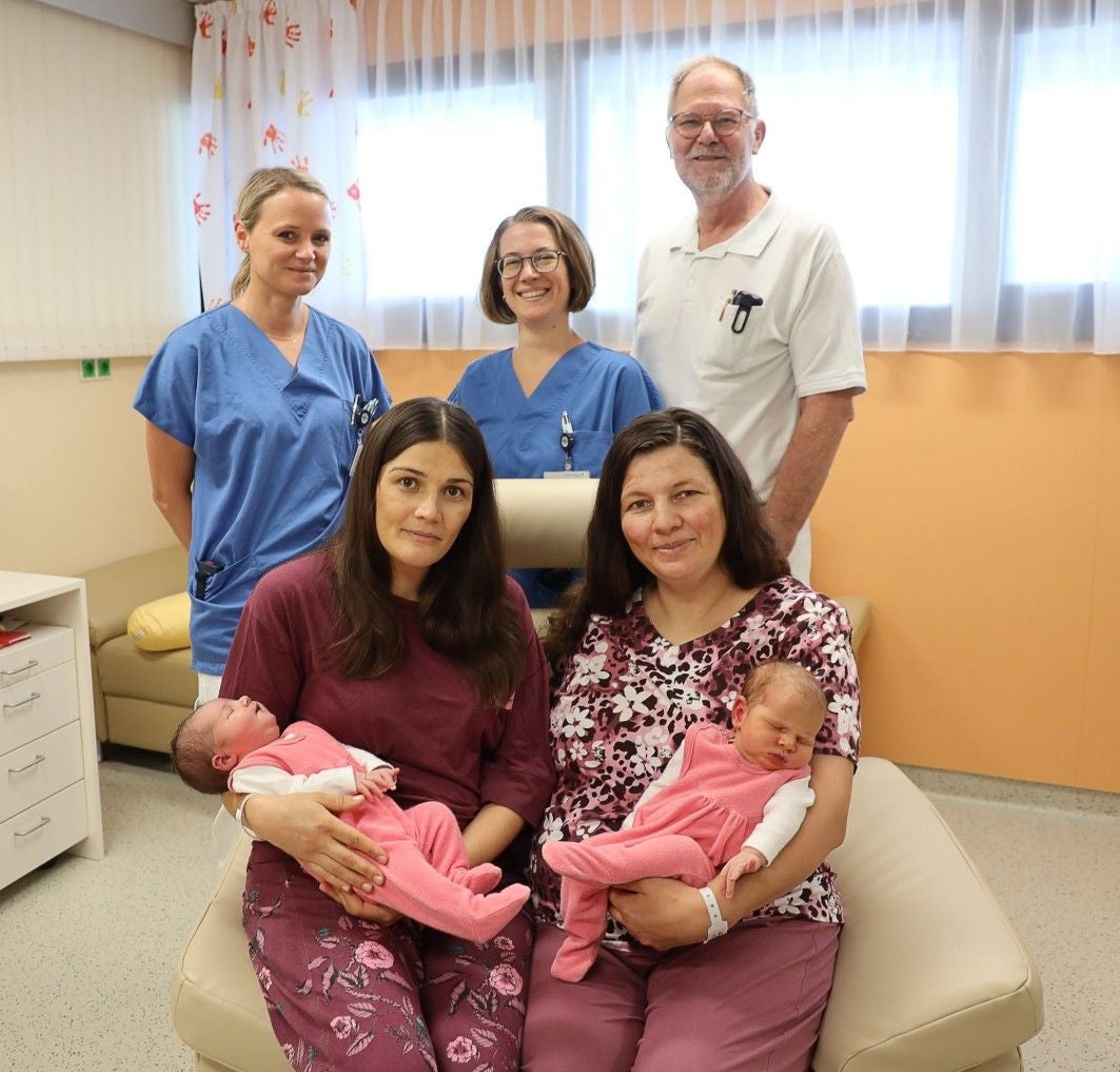 Zwei Frauen mit Babys auf dem Schoß, drei Krankenhausmitarbeiter im Hintergrund.