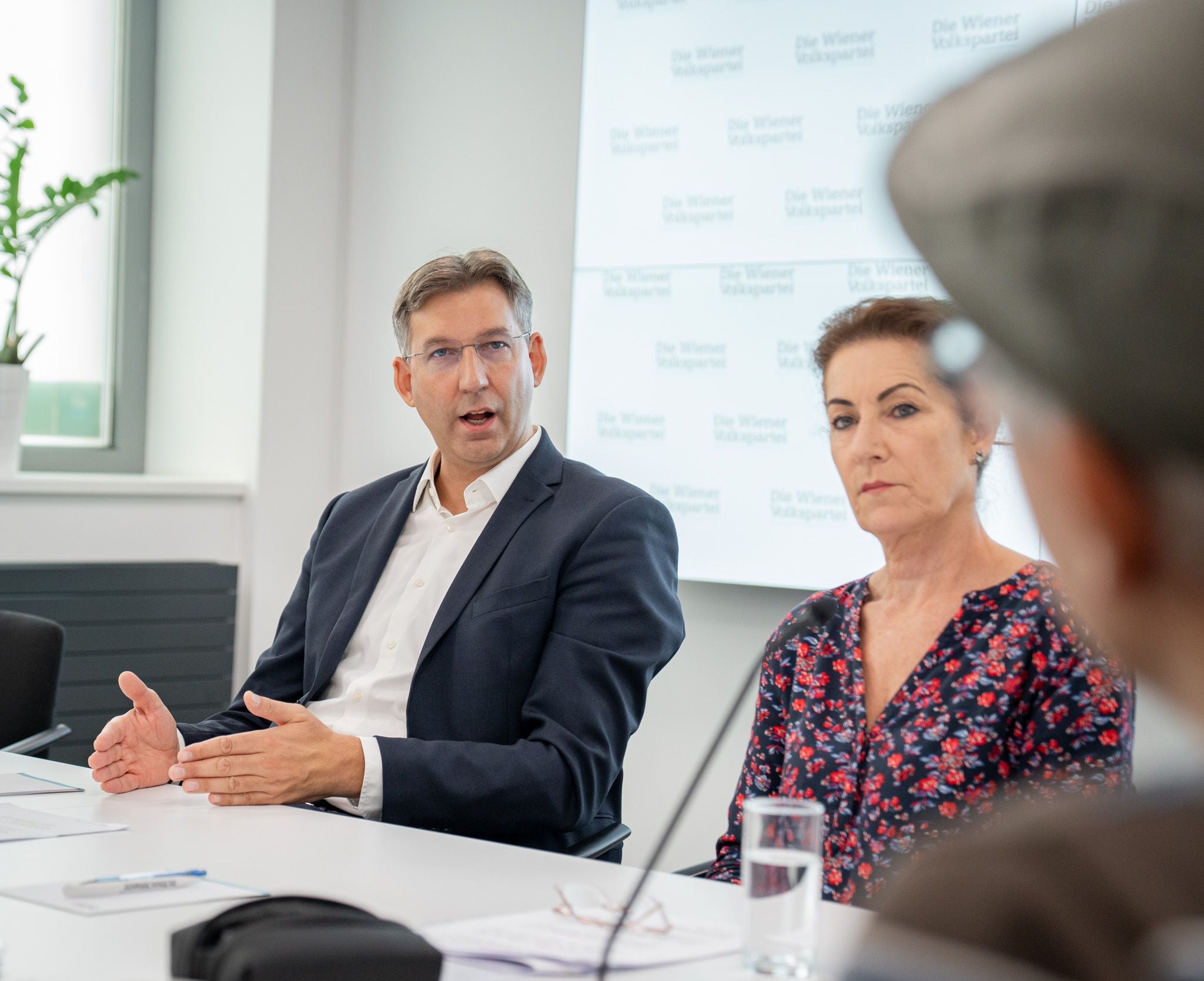Ein Mann und eine Frau sitzen bei einer Besprechung an einem Tisch mit Mikrofonen und Notizen.