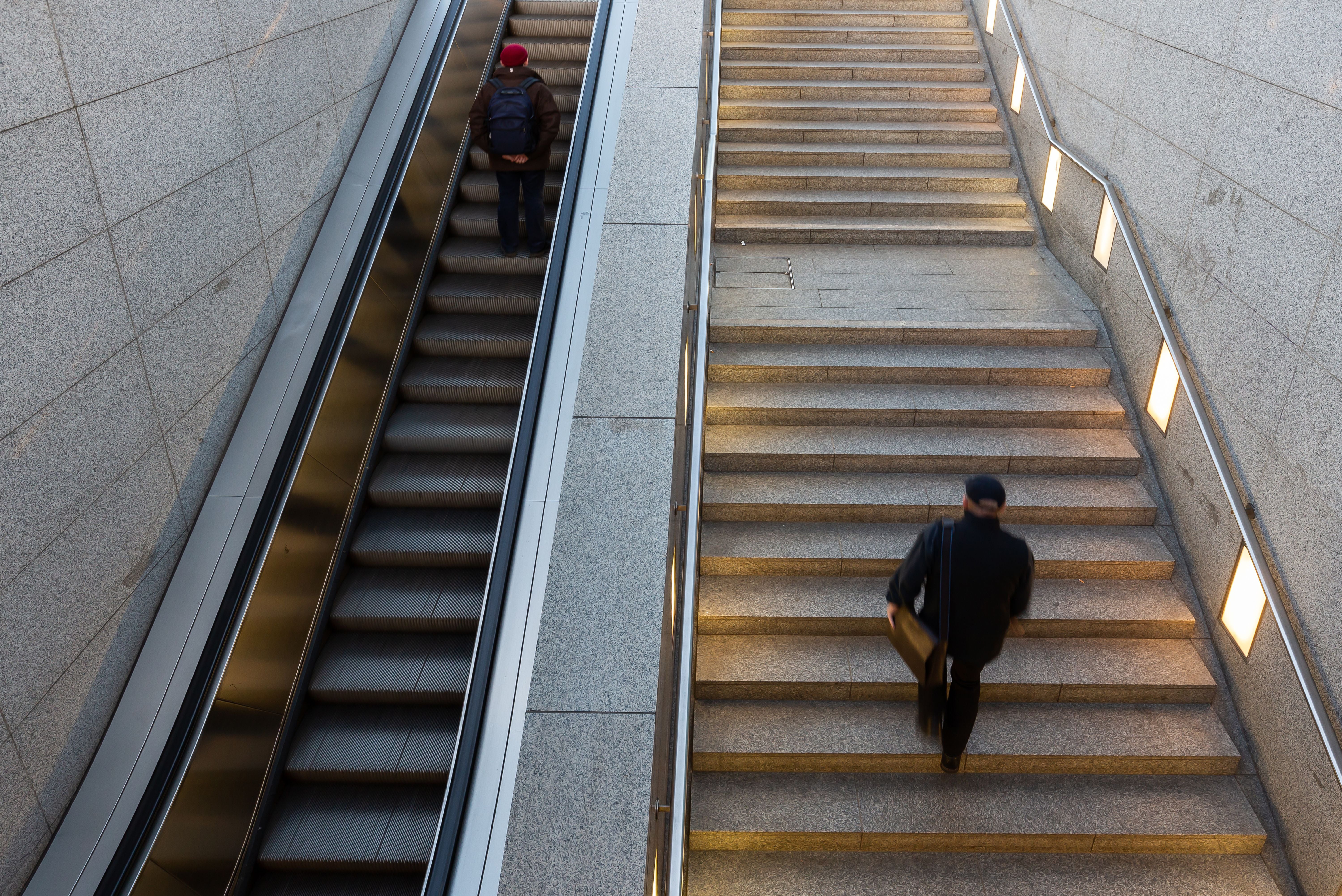 Eine Person geht die Treppe hinauf, eine andere fährt mit der Rolltreppe nach unten.