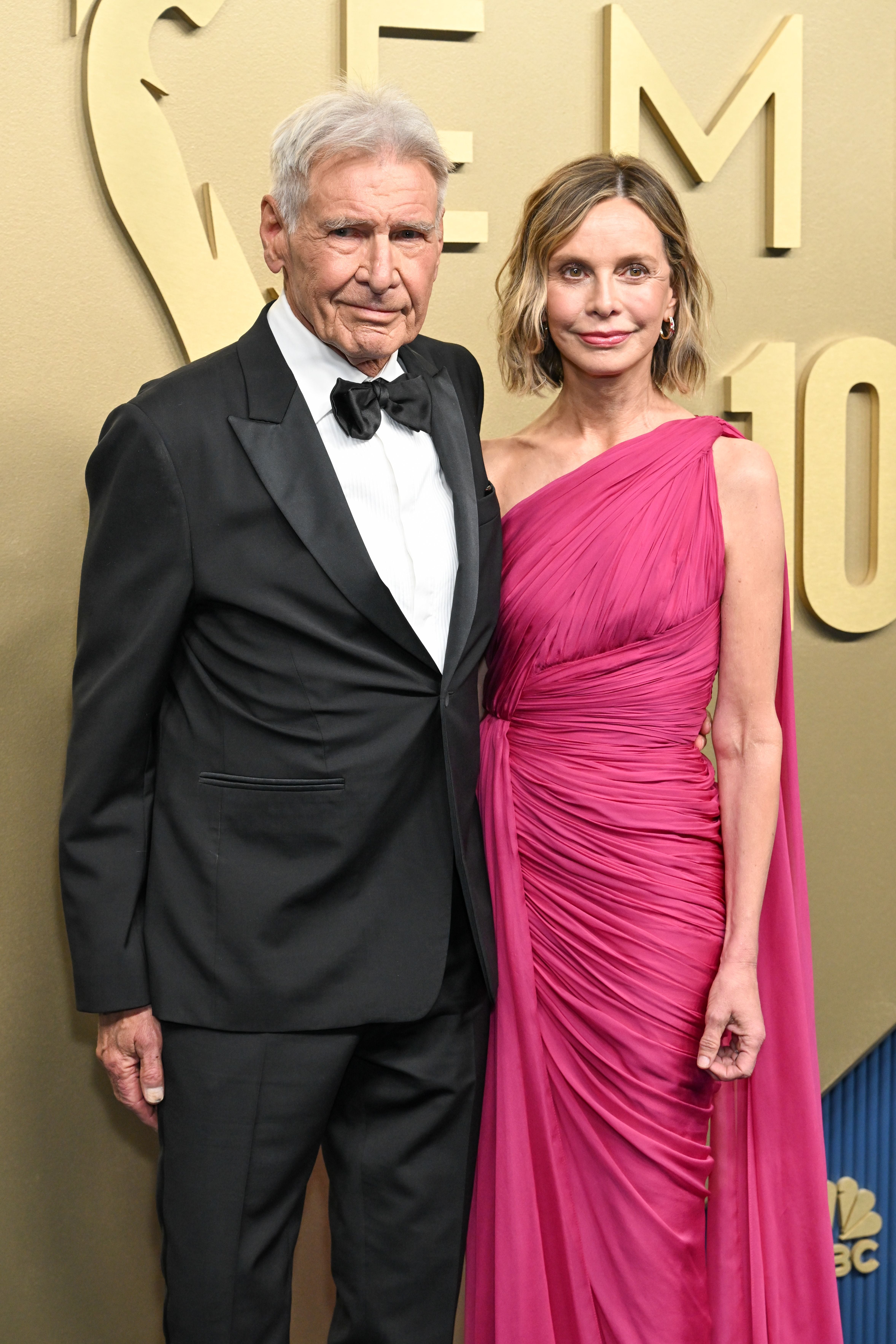 Ein Mann im Smoking und eine Frau im pinken Abendkleid posieren vor einer goldenen Wand.