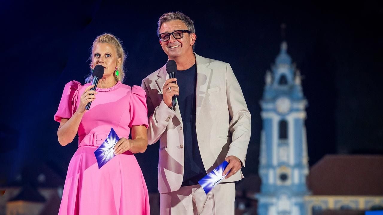 Eine Frau im pinken Kleid und ein Mann im beigen Anzug moderieren auf einer Bühne mit Mikrofonen.