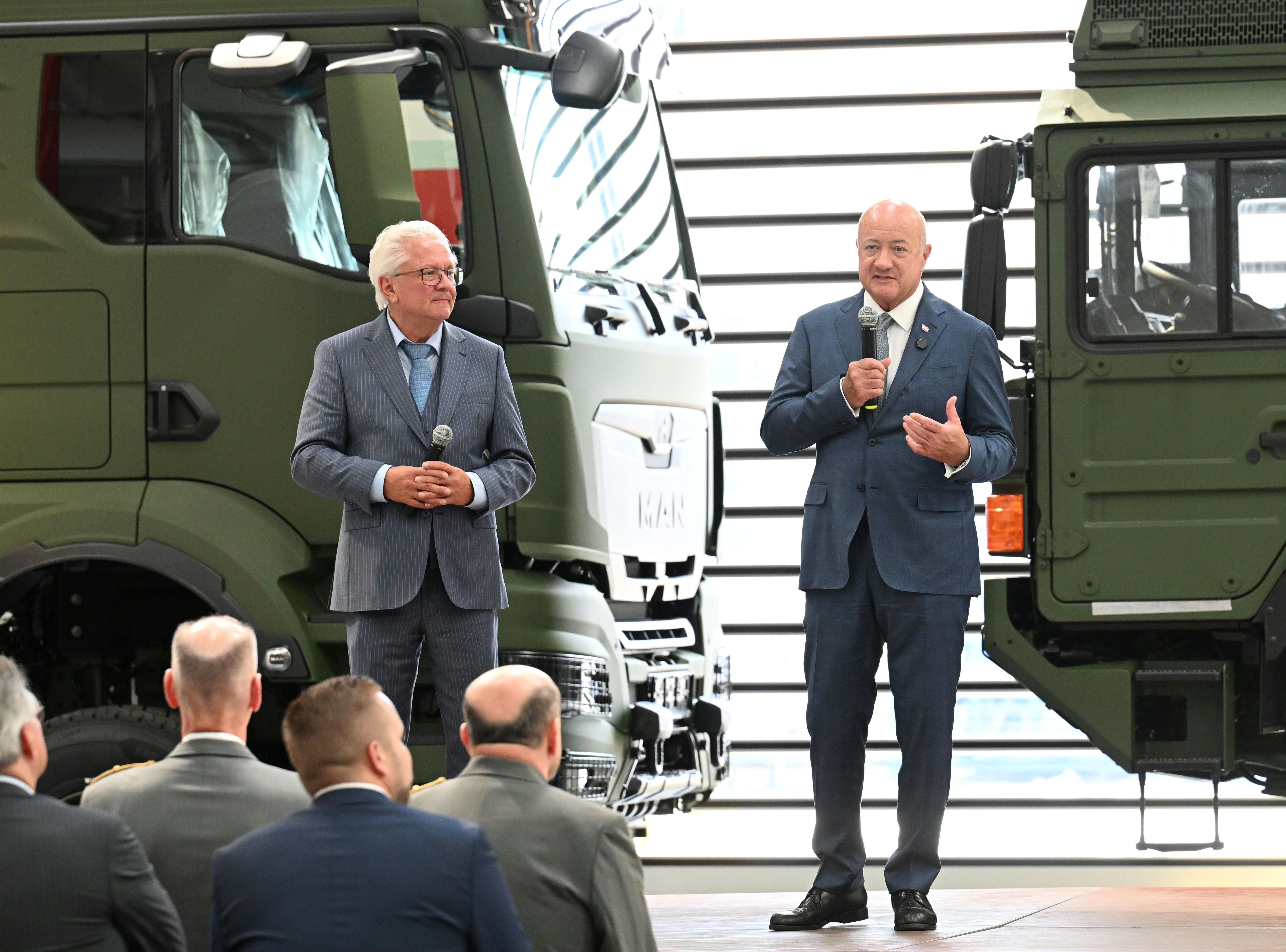 Zwei Männer im Anzug sprechen mit Mikrofonen vor Militärfahrzeugen auf einer Bühne.