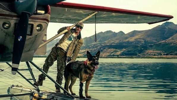 Mann mit Hund steht auf einem Wasserflugzeug vor einer Bergkulisse am See.