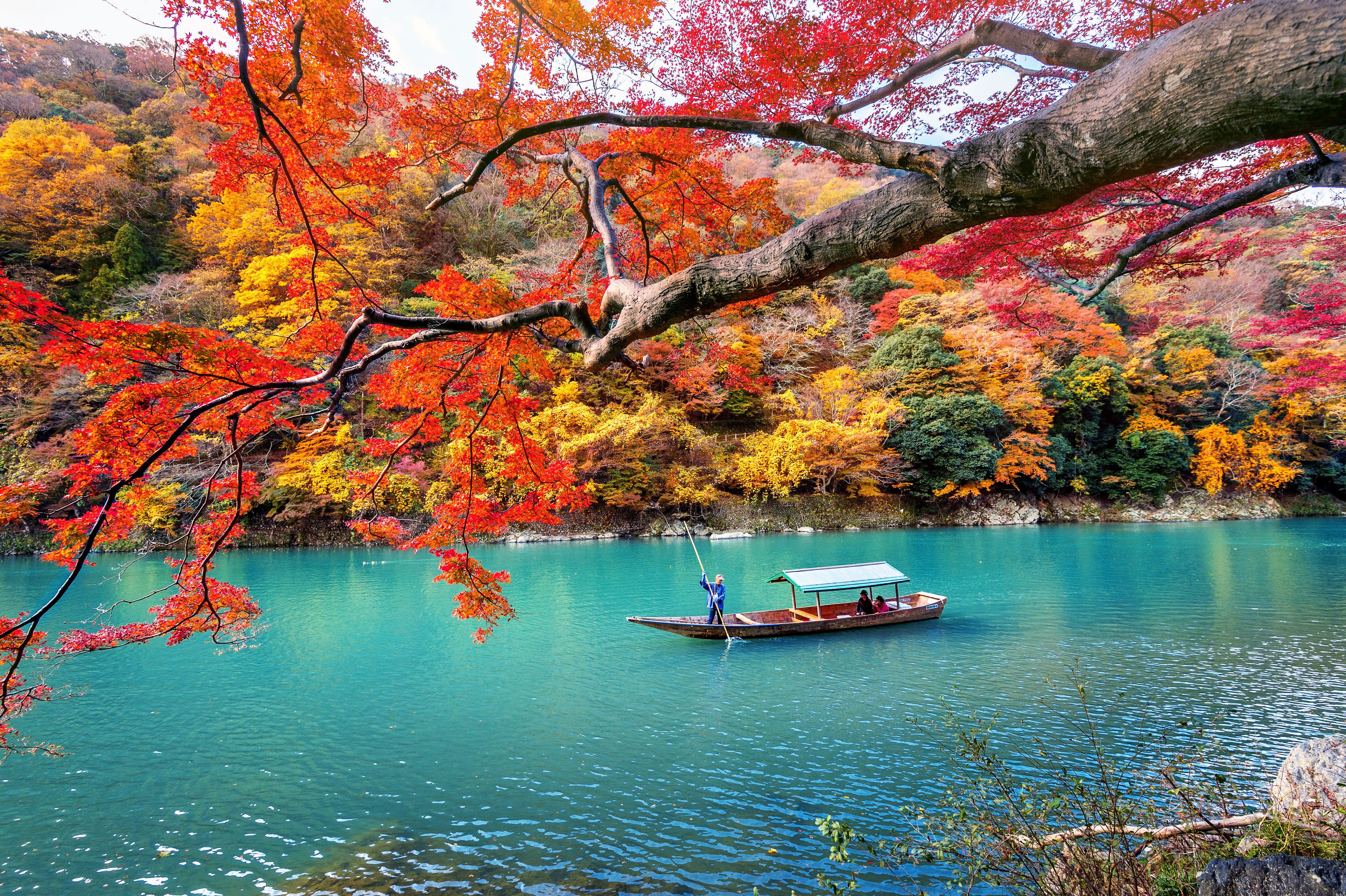 Boot auf türkisblauem Fluss, umgeben von bunten Herbstbäumen in Arashiyama, Kyoto.