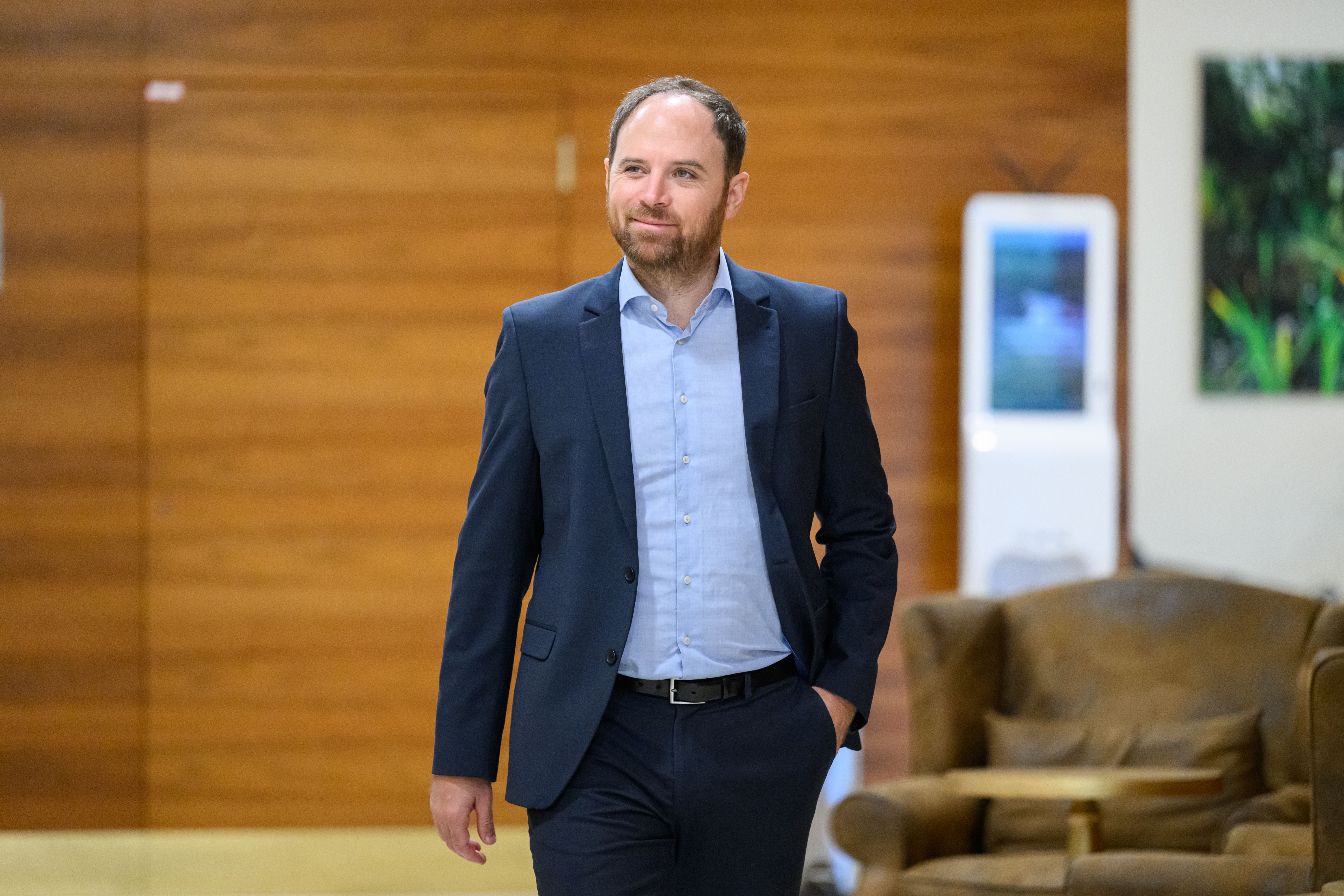 Ein Mann im Anzug geht durch einen Raum mit brauner Wand und Ledersessel im Hintergrund.