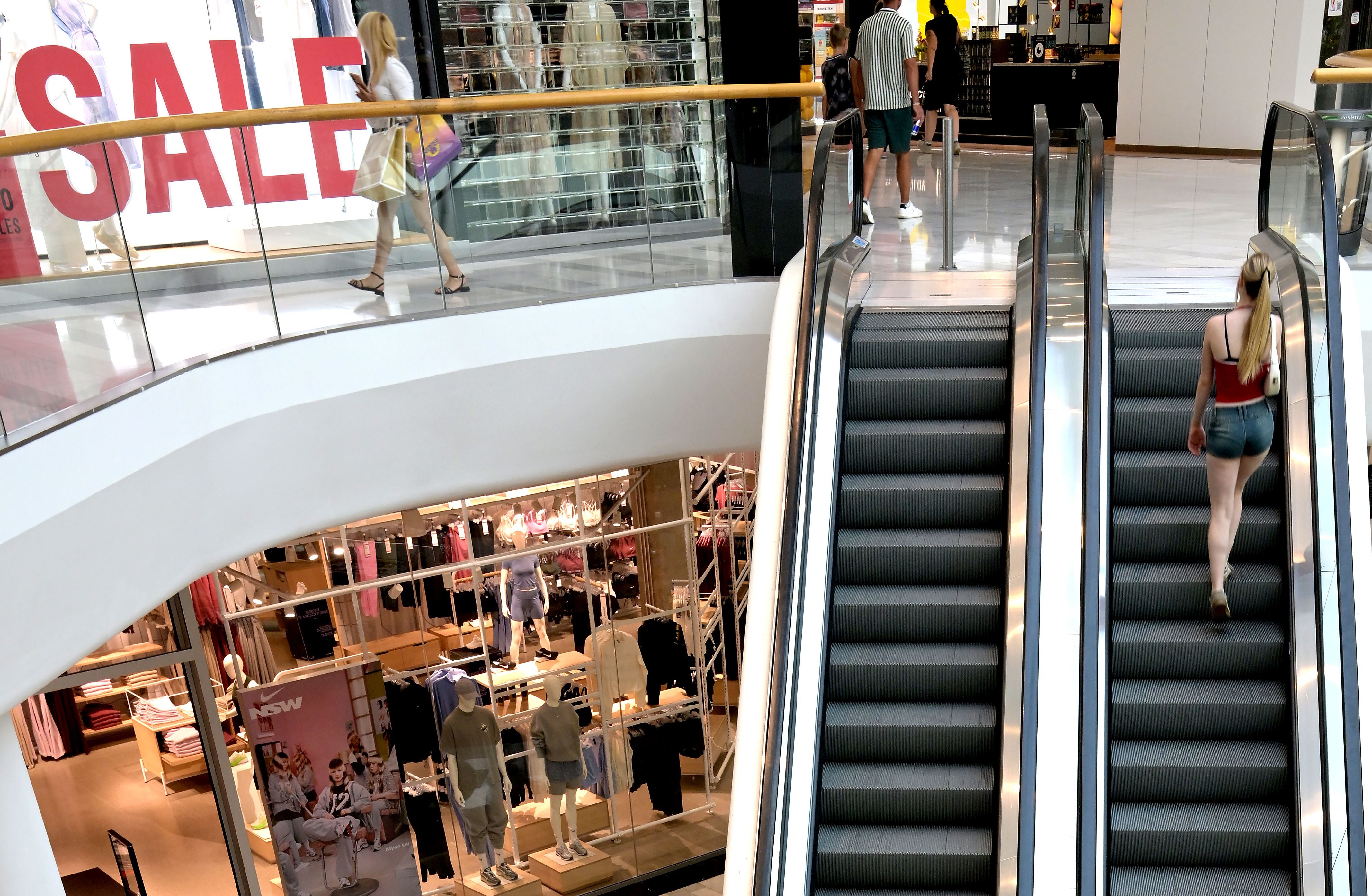 Frau fährt Rolltreppe im Einkaufszentrum, große SALE-Werbung und Schaufenster im Hintergrund.