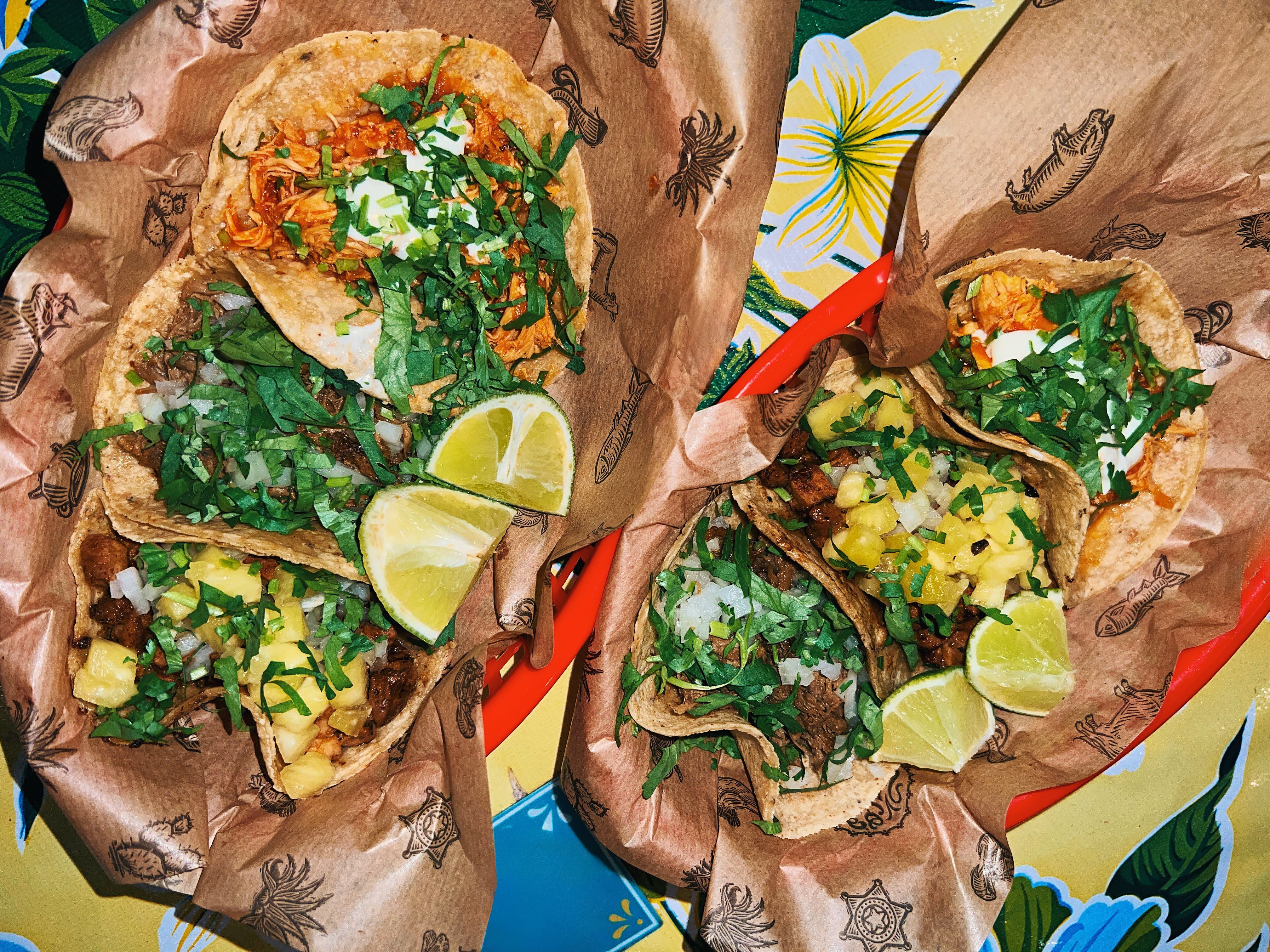 Zwei Körbe mit verschiedenen Tacos, Limettenspalten und Kräutern auf buntem Untergrund.