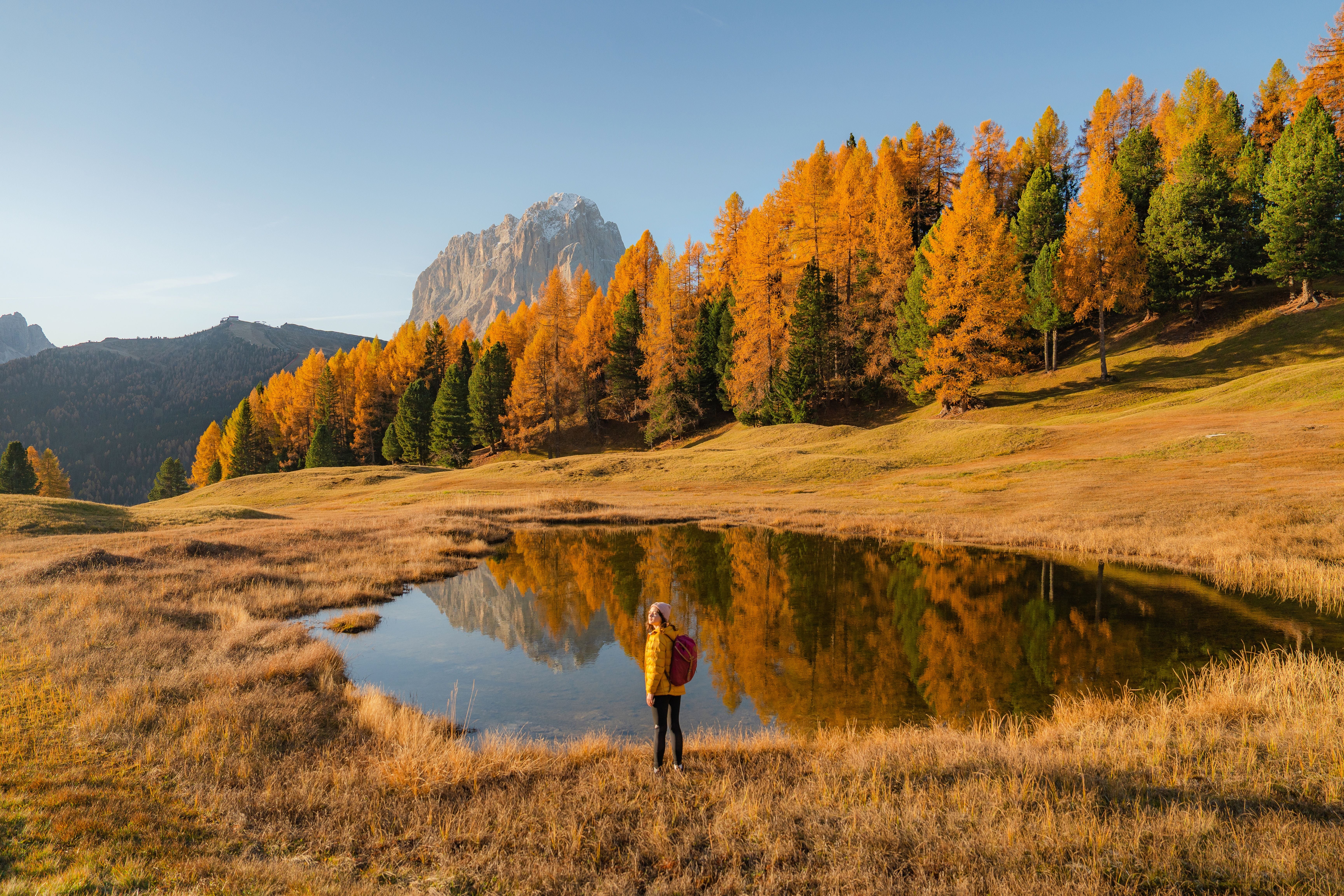 Frau in gelber Jacke steht an einem kleinen See vor herbstlichen Bäumen in den Dolomiten.