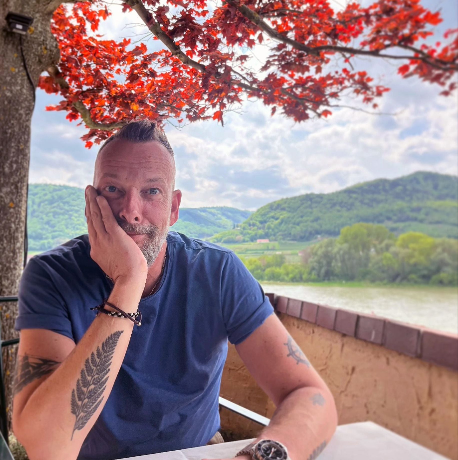 Mann sitzt draußen unter rotem Baum mit Blick auf grüne Hügel und Fluss im Hintergrund.