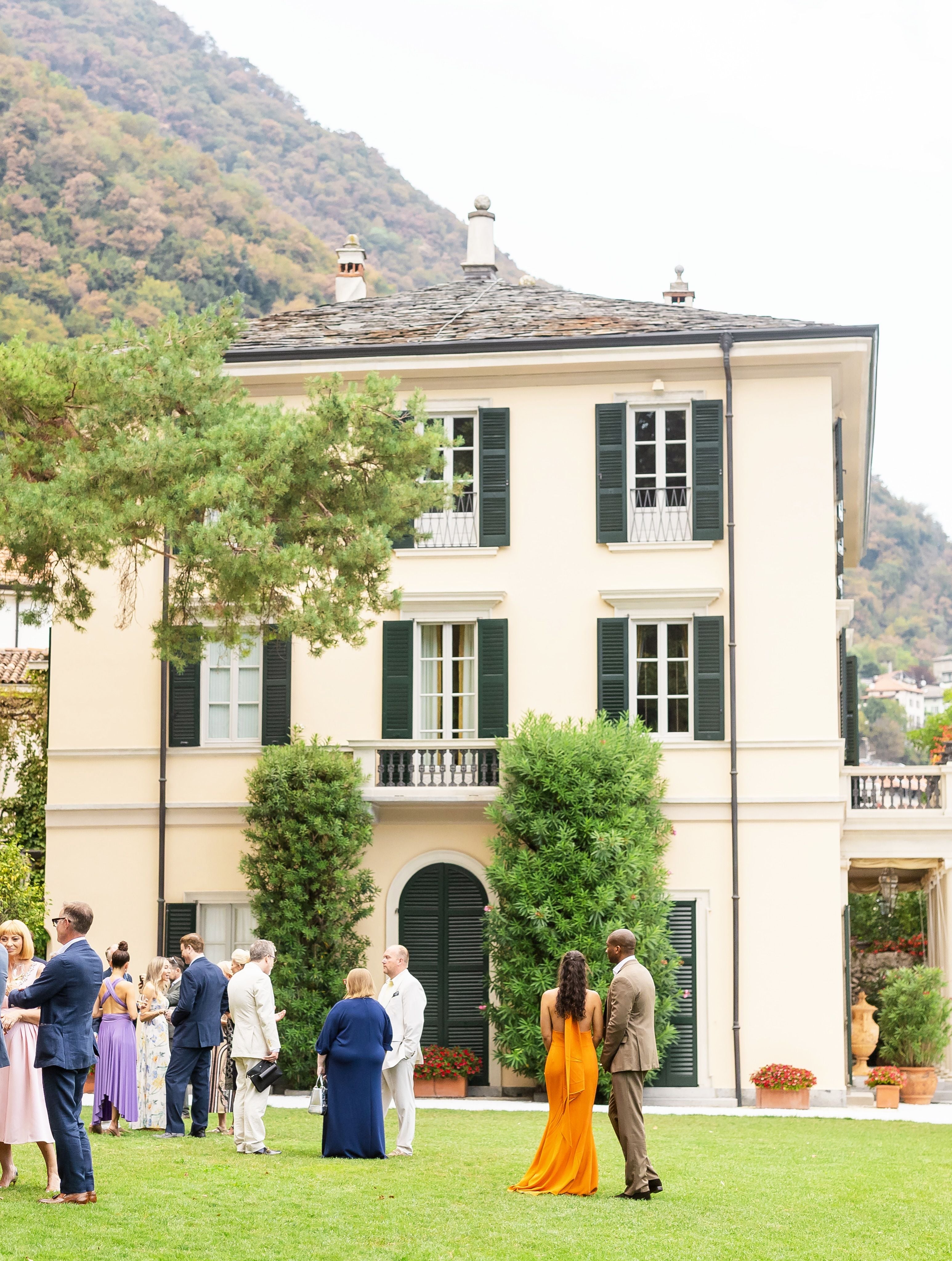 Mehrere elegant gekleidete Menschen stehen auf einer Wiese vor einer großen Villa mit Bergkulisse.
