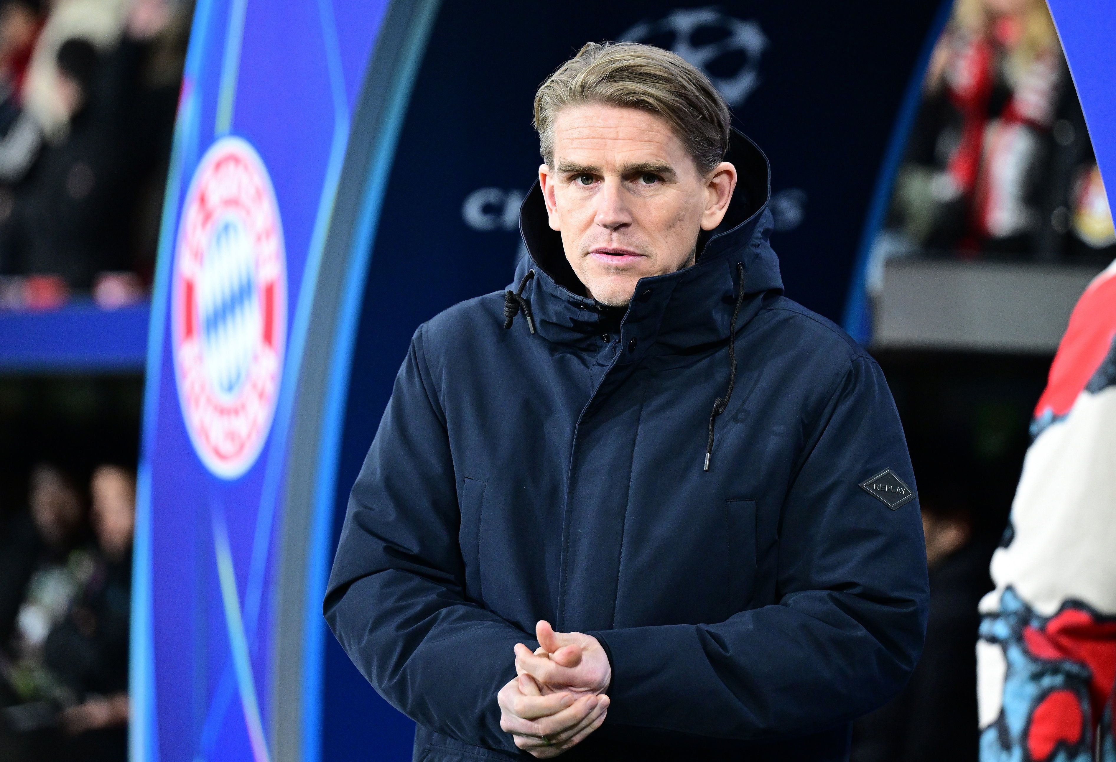 Ein Mann im dunklen Mantel steht vor Bayern-Logo während eines Fußballspiels.