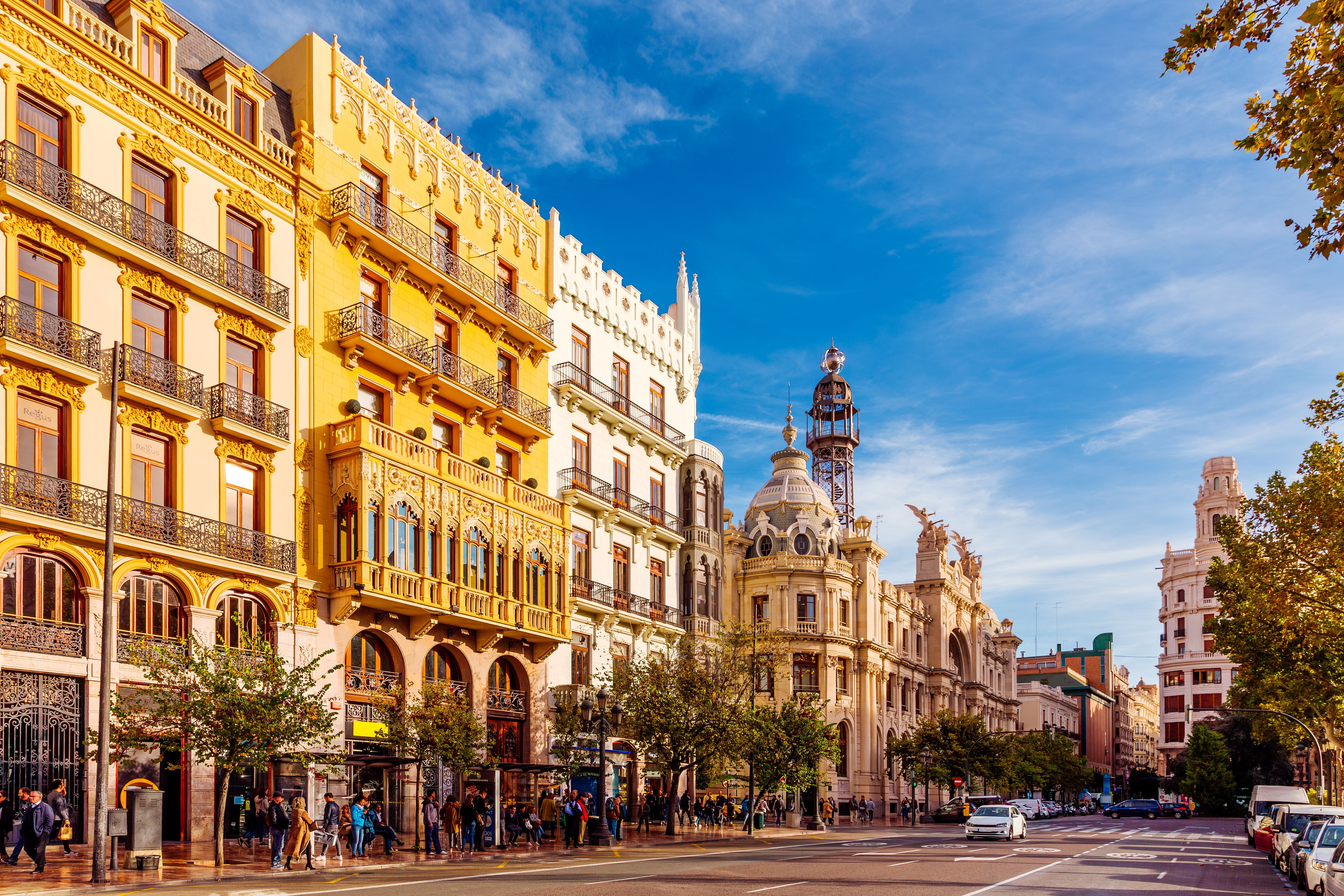 Prächtige historische Gebäude an einer sonnigen Straße in Valencia mit Fußgängern und Autos.