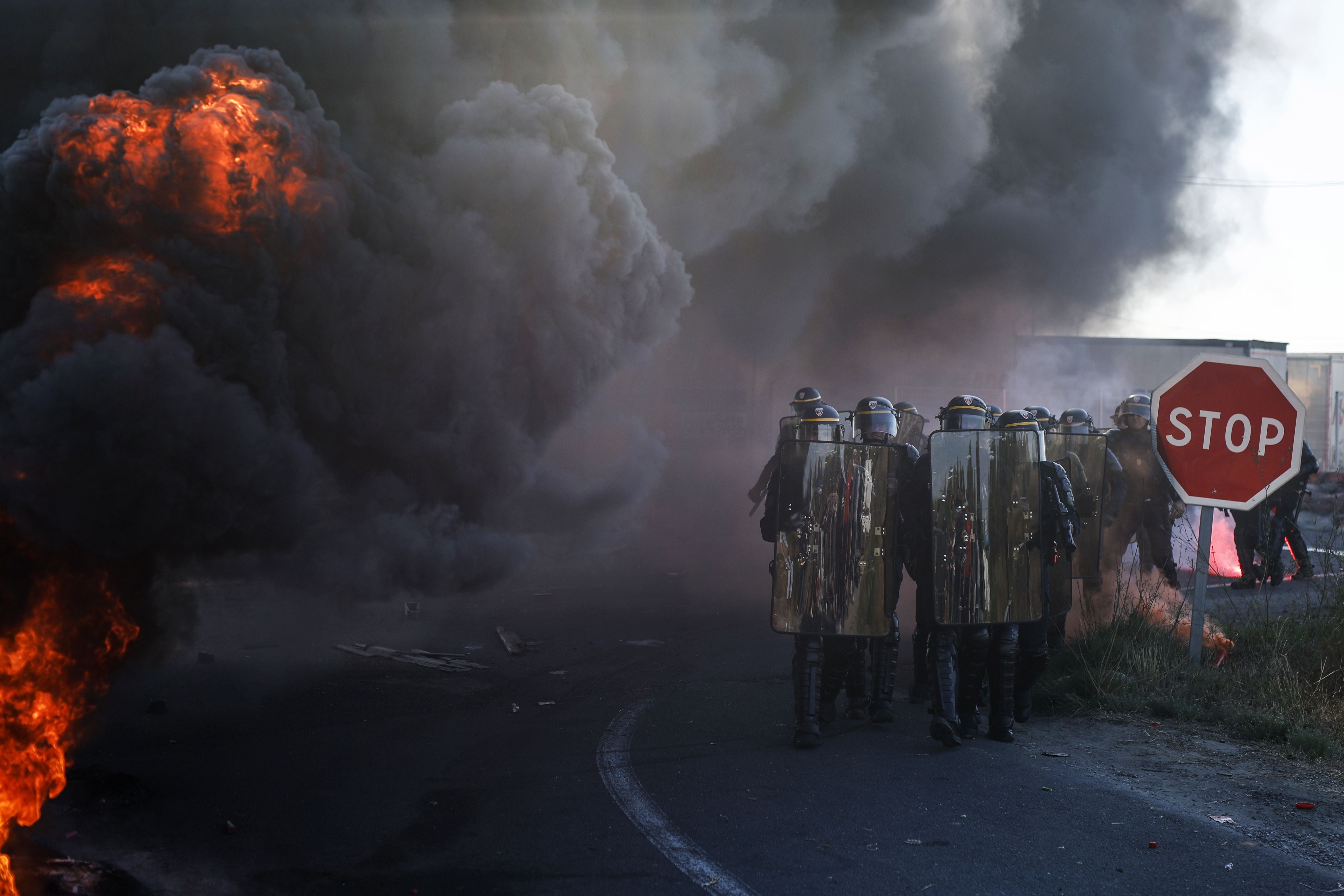 Polizisten mit Schilden stehen nahe eines Feuers und dichter Rauch bei einem Streik mit STOP-Schild.
