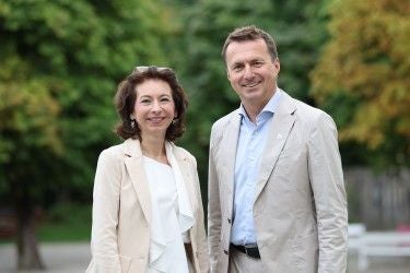 Eine Frau und ein Mann stehen im Freien und lächeln in die Kamera, Bäume im Hintergrund.