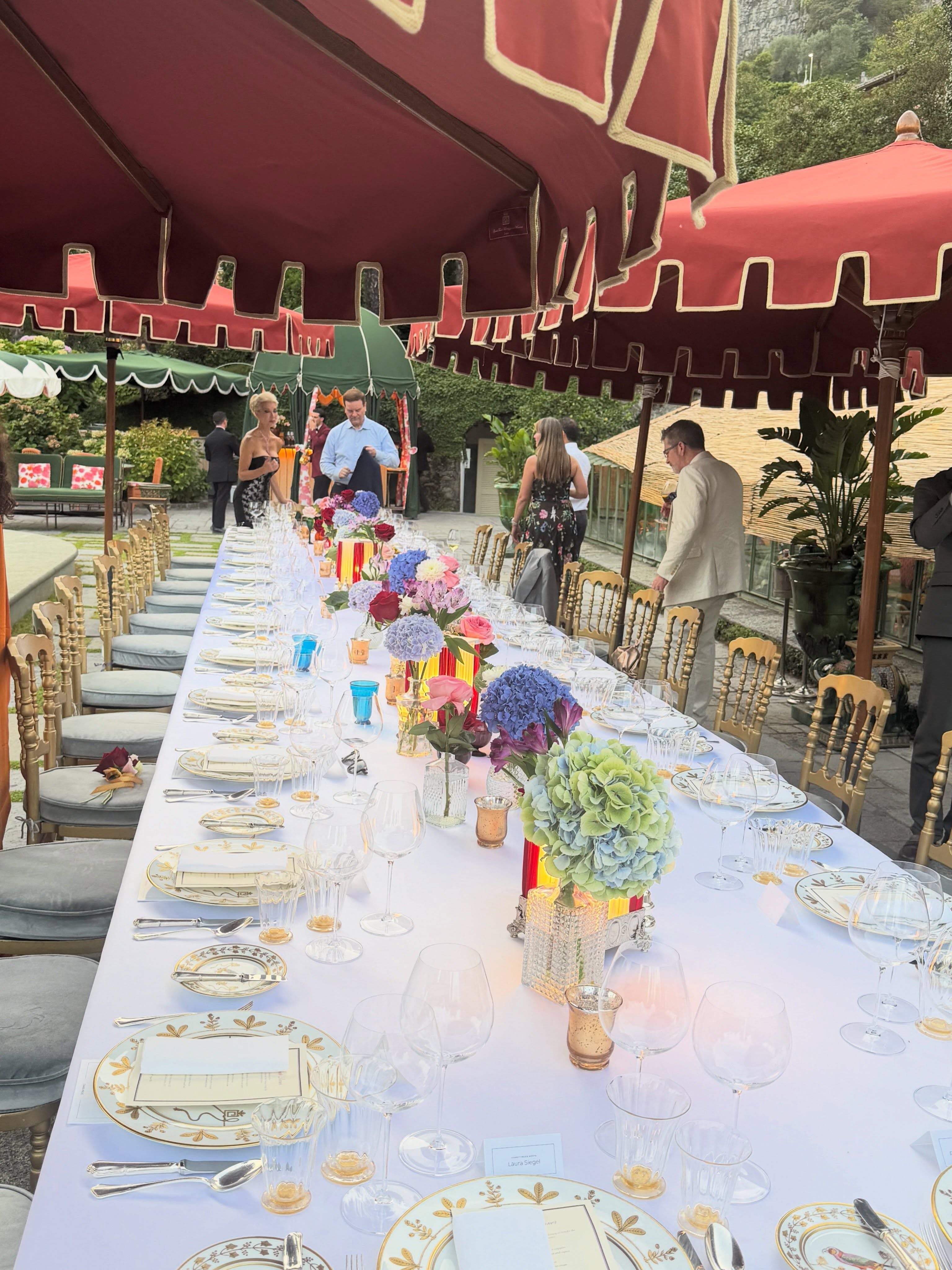Lange, elegant gedeckte Festtafel im Freien mit Blumen und festlich gekleideten Gästen.