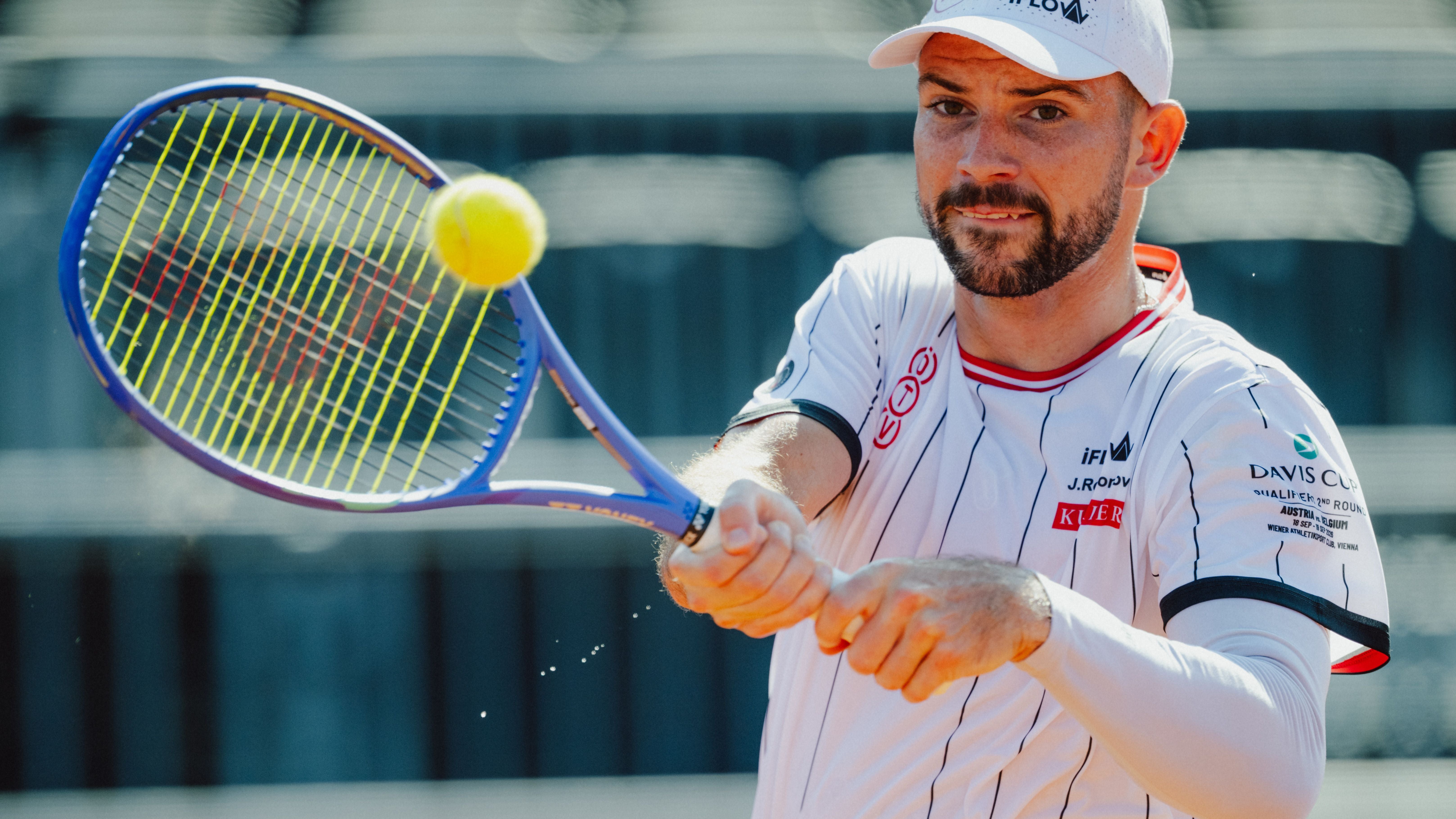 Jurij Rodionov im weißen ÖTV-Outfit holt mit der Rückhand zum Schlag aus, während der Tennisball nahe am Schlägerrahmen fliegt.