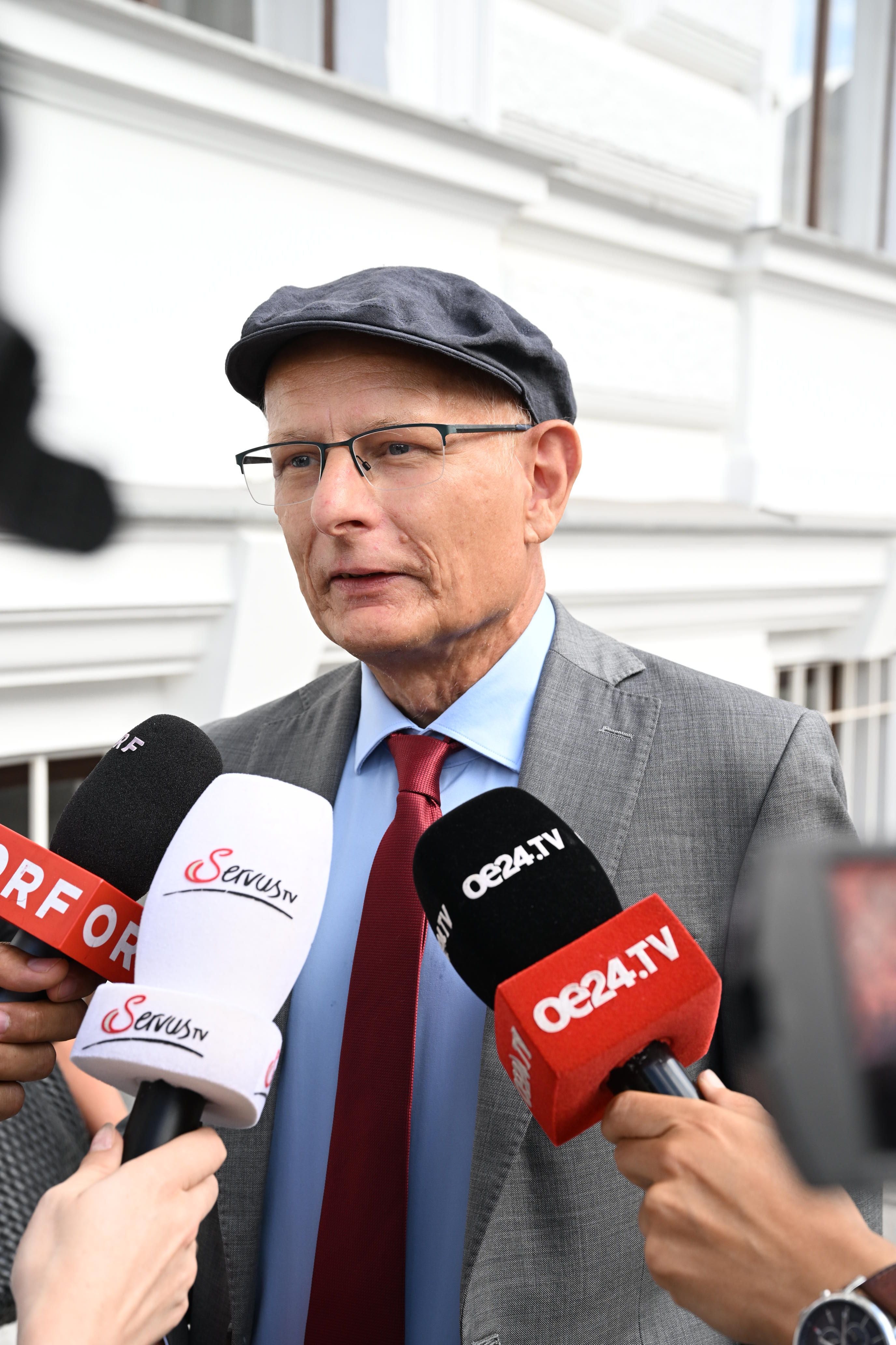 Mann mit Kappe wird draußen von mehreren Mikrofonen verschiedener TV-Sender interviewt.