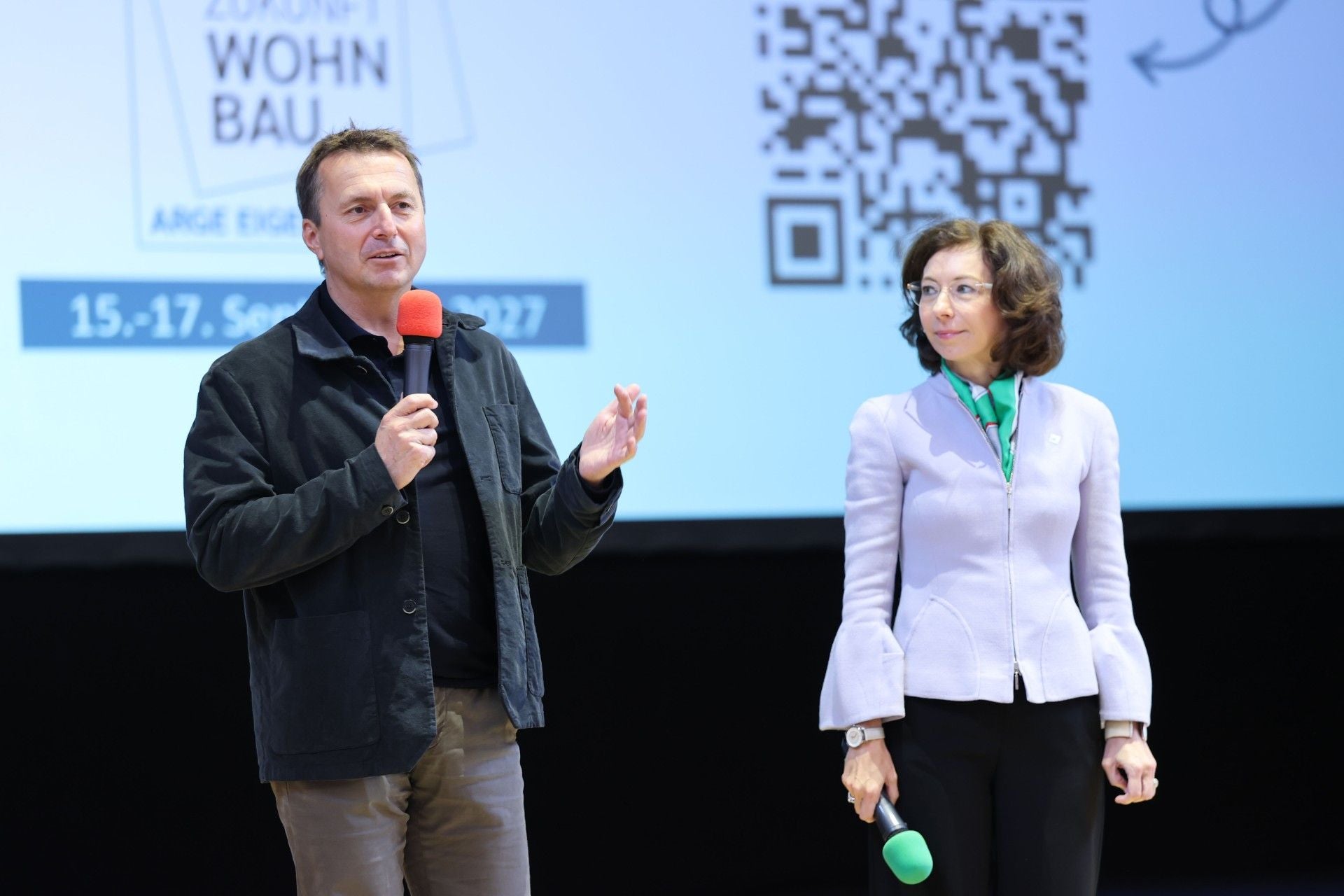 Ein Mann und eine Frau sprechen mit Mikrofonen vor einer Präsentationswand bei einer Tagung.