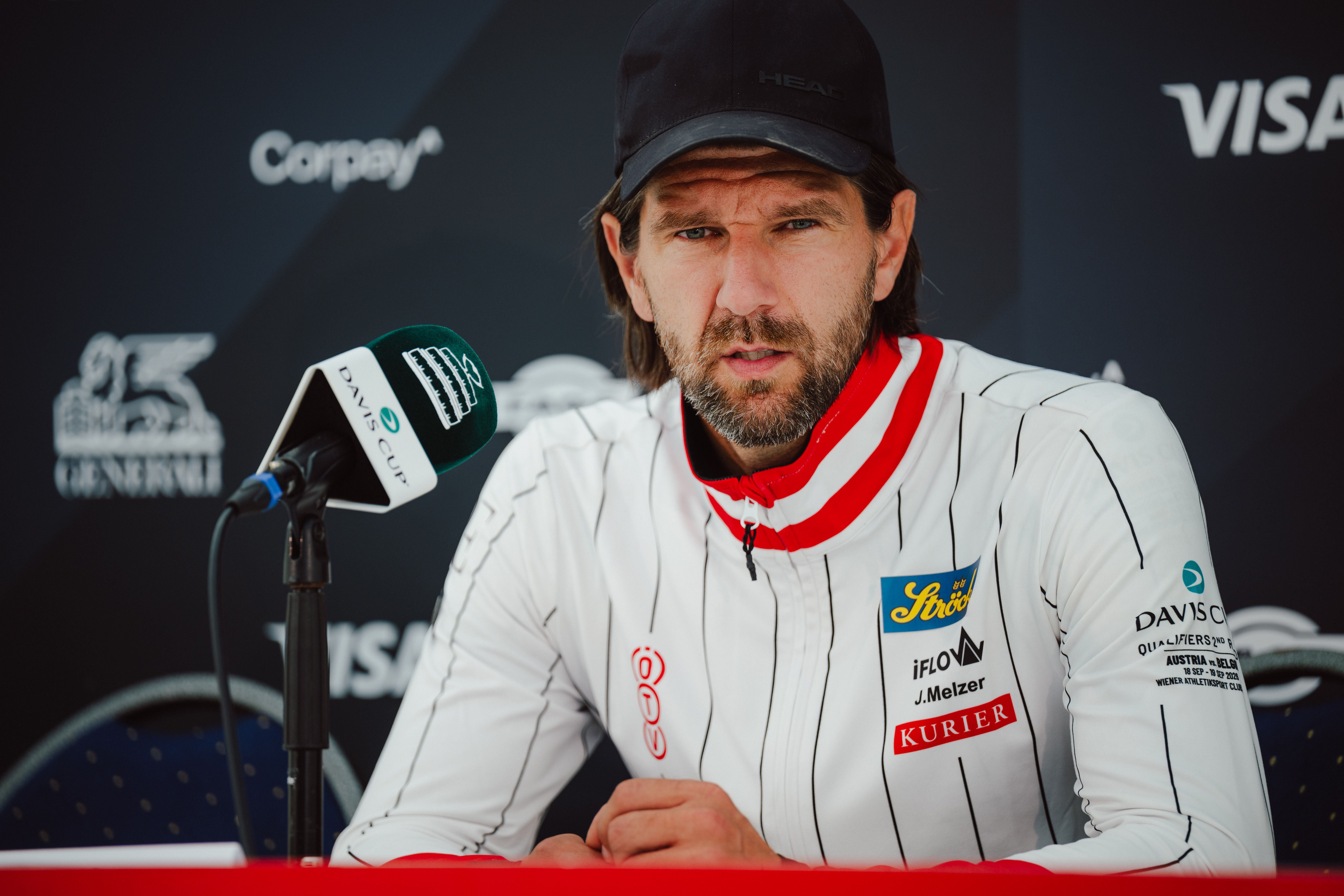 ÖTV-Kapitän Jürgen Melzer im weißen Trainingsauftakt-Kappen-Look blickt bei der Pressekonferenz fokussiert hinter einem Mikrofon in die Kamera.