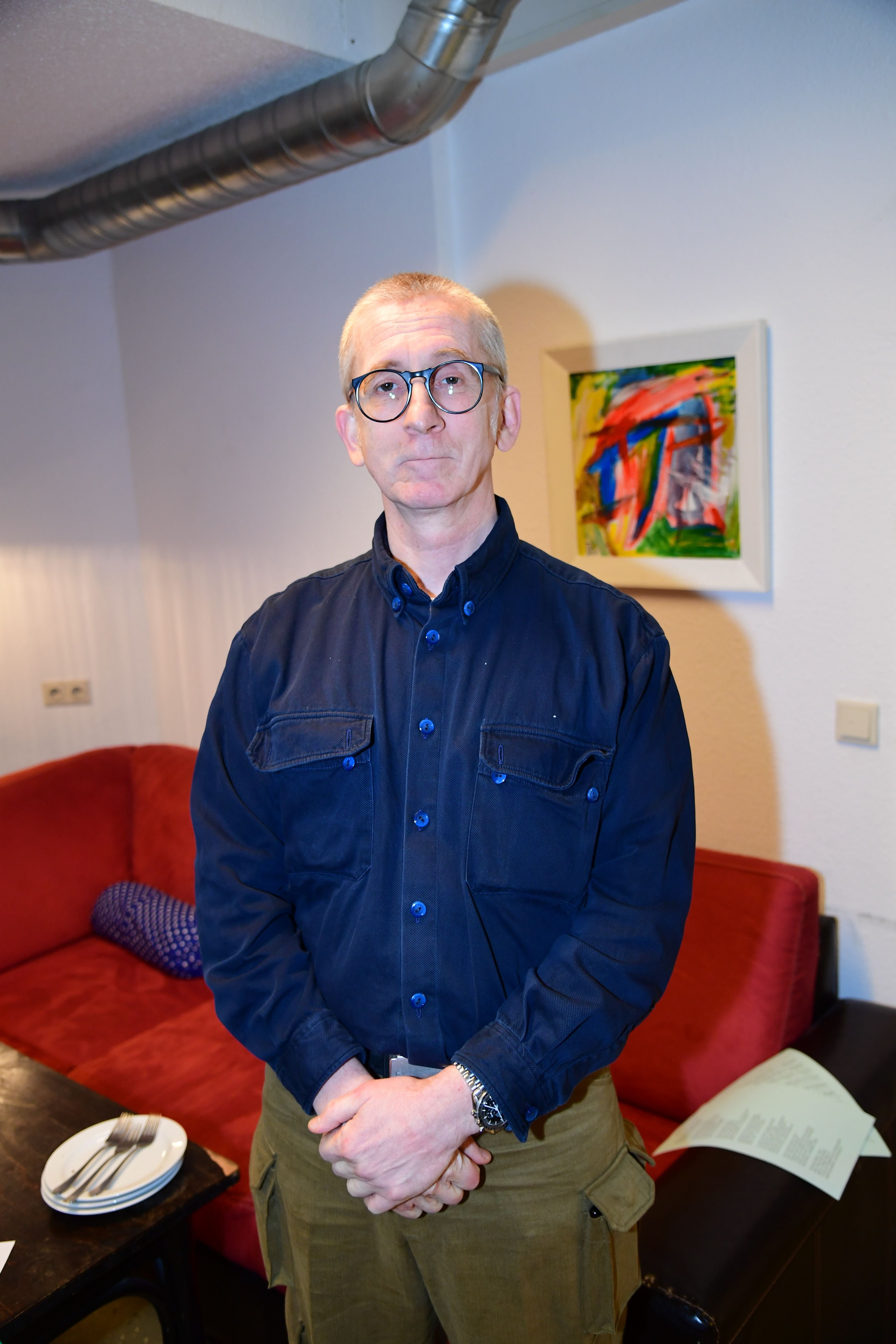 Ein Mann mit Brille steht vor einem roten Sofa in einem modern eingerichteten Raum.