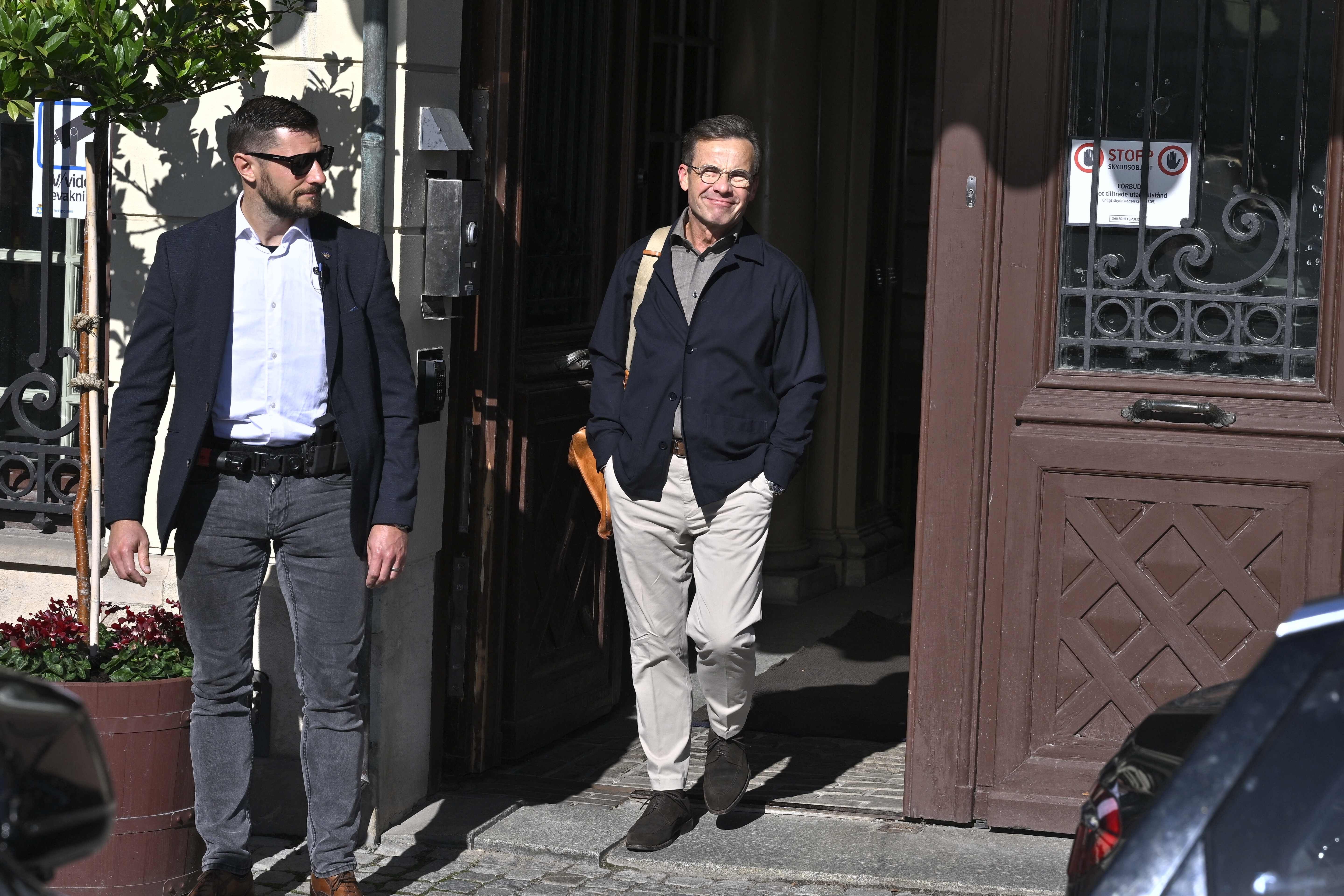 Zwei Männer verlassen ein Gebäude bei Sonnenschein, einer mit Umhängetasche und Brille.