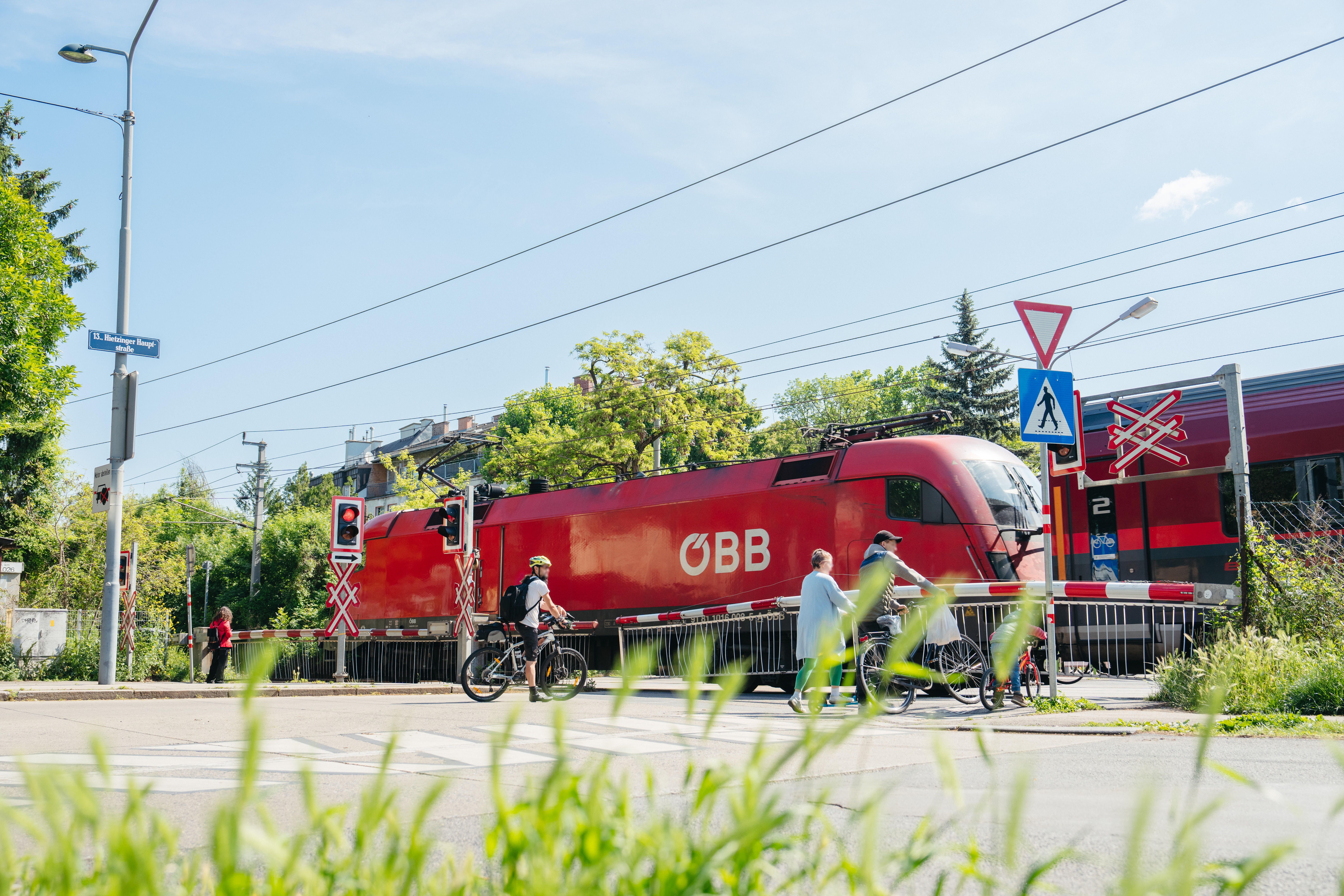 Roter ÖBB-Zug fährt an geschlossener Schranke vorbei; Menschen warten mit Fahrrädern am Bahnübergang.