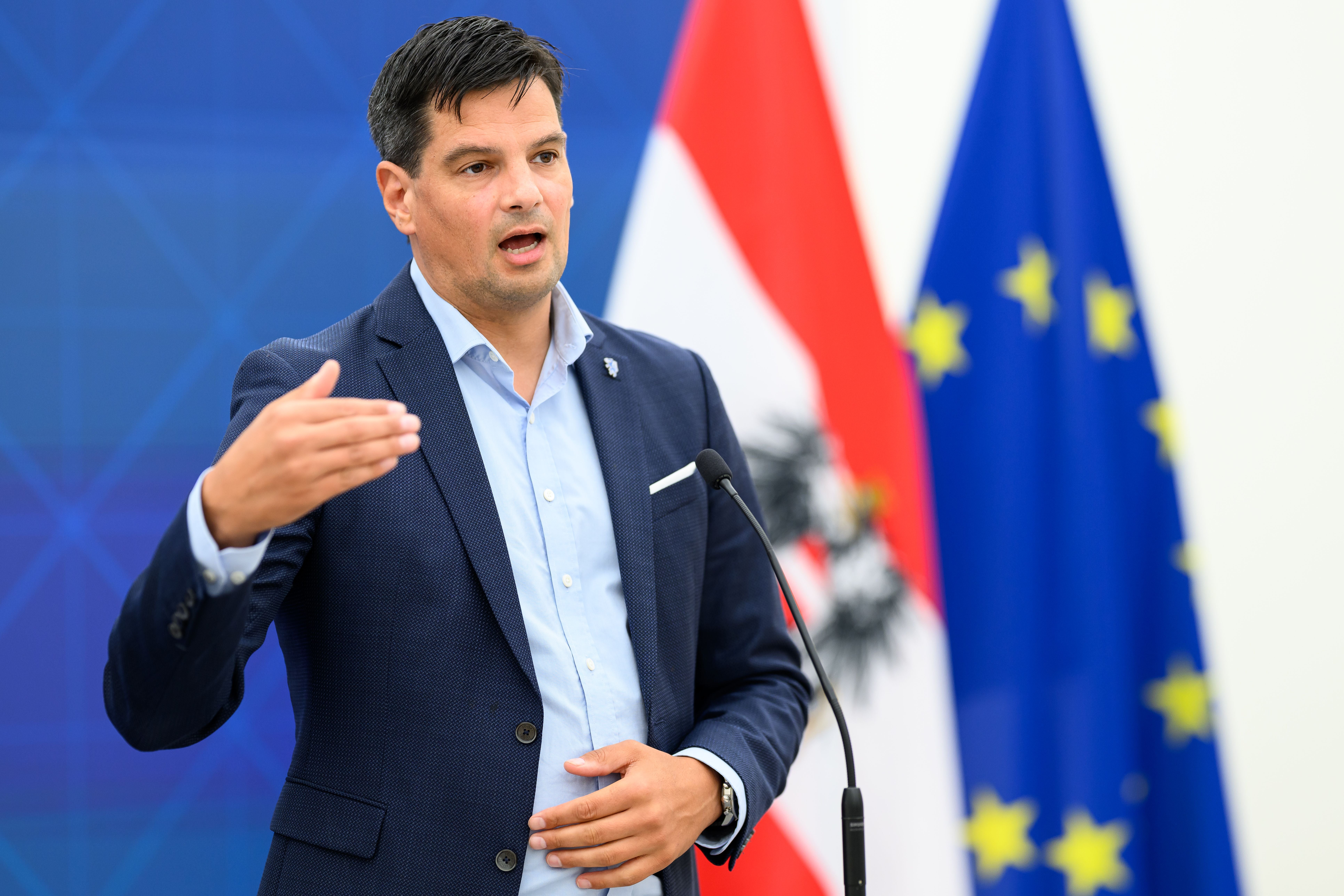 Ein Mann spricht bei einer Pressekonferenz vor österreichischer und EU-Flagge am Mikrofon.