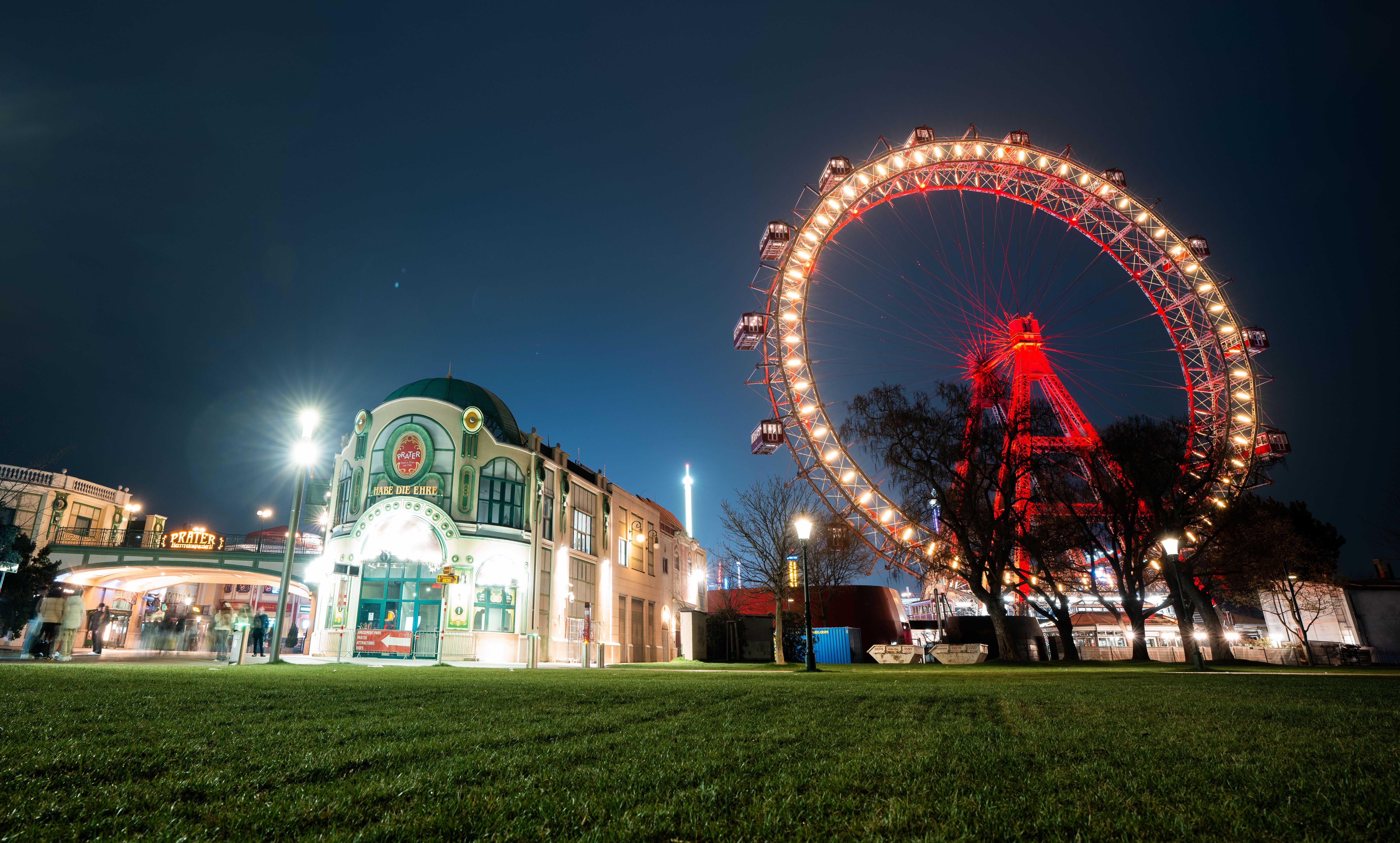 Beleuchtetes Riesenrad und Gebäude im Wiener Prater bei Nacht, mit Wiese im Vordergrund.