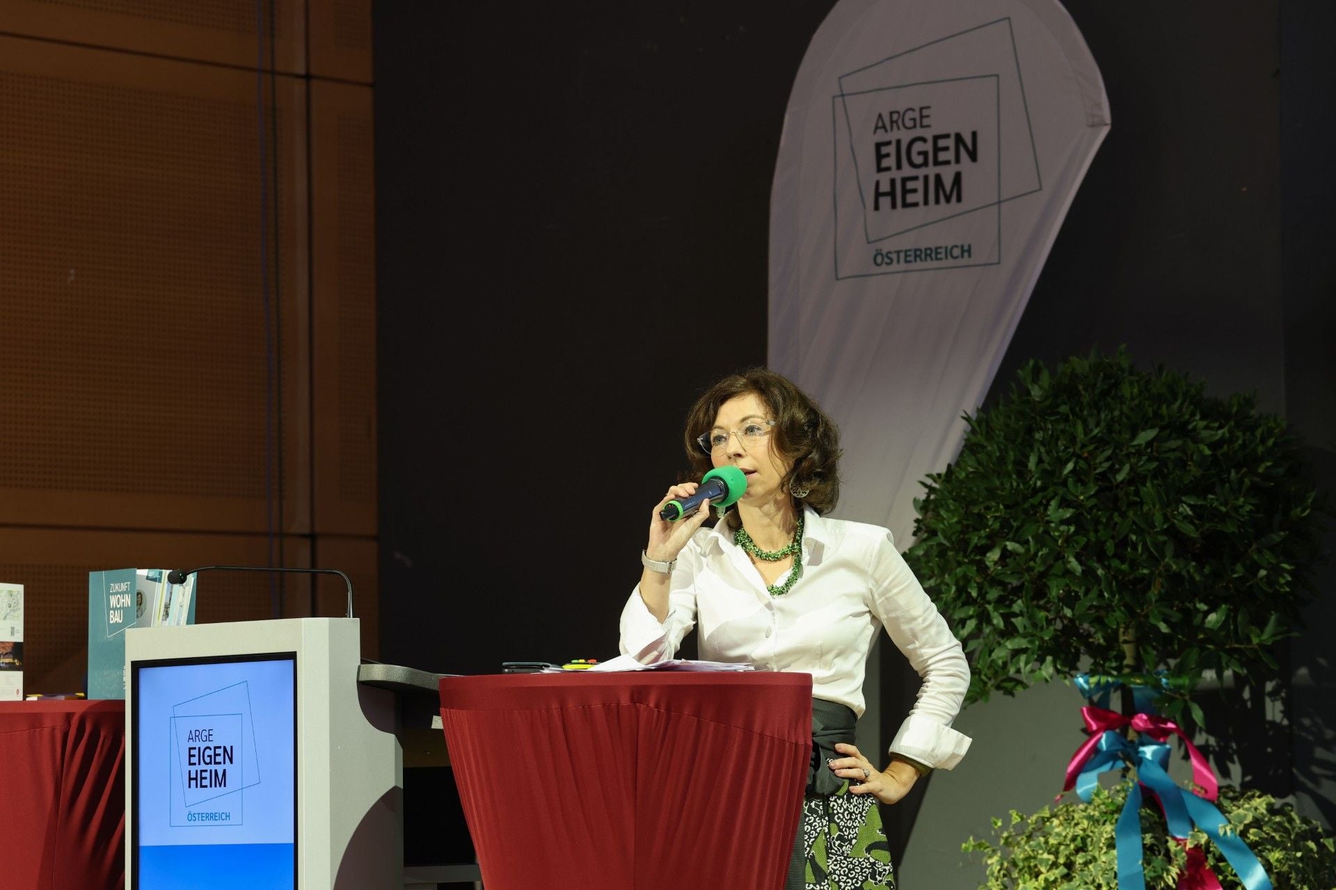 Frau spricht bei einer Veranstaltung vor Mikrofon, im Hintergrund Banner von ARGE EIGENHEIM.