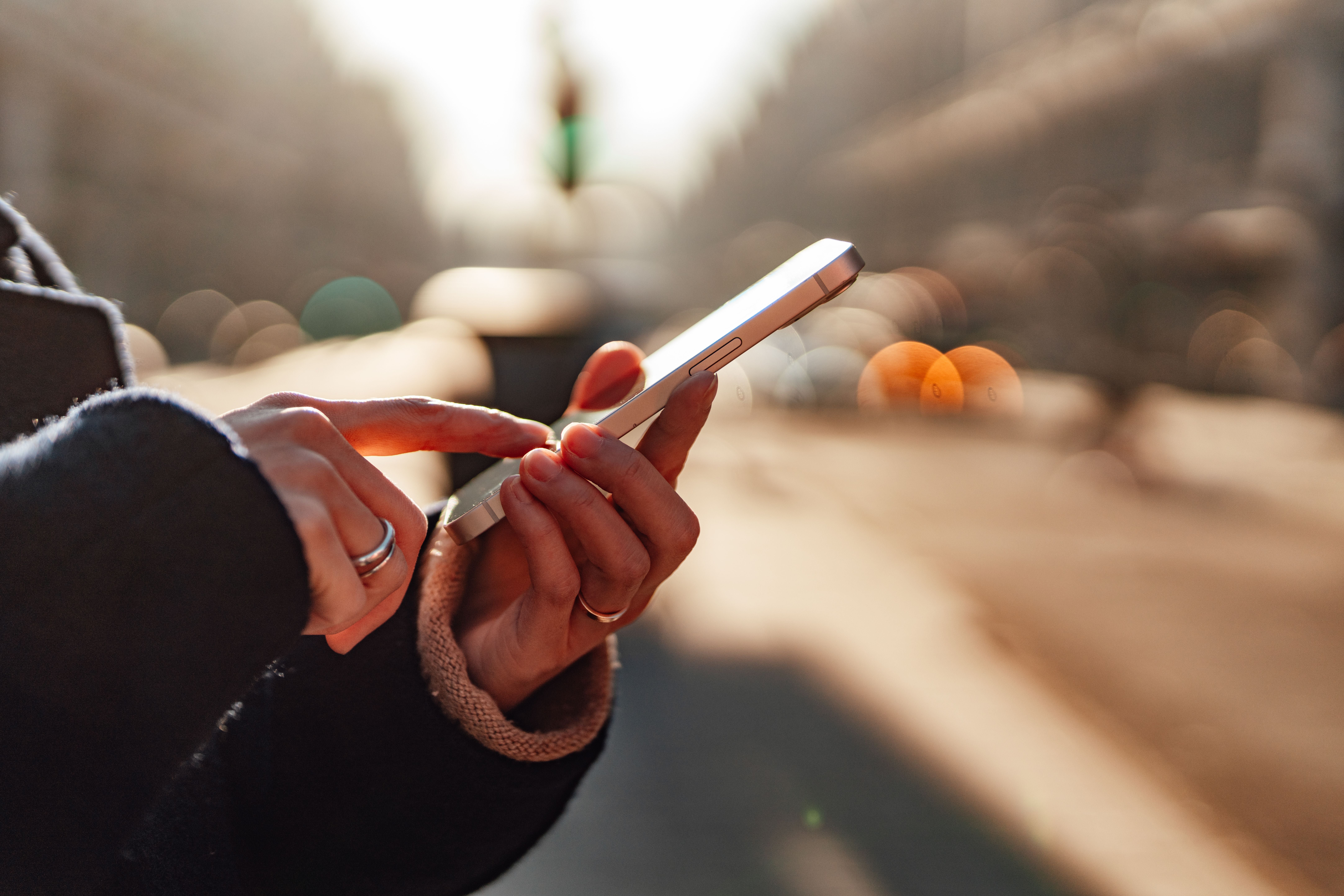 Frauenhände bedienen ein Smartphone auf einer sonnigen, unscharfen Stadtstraße.