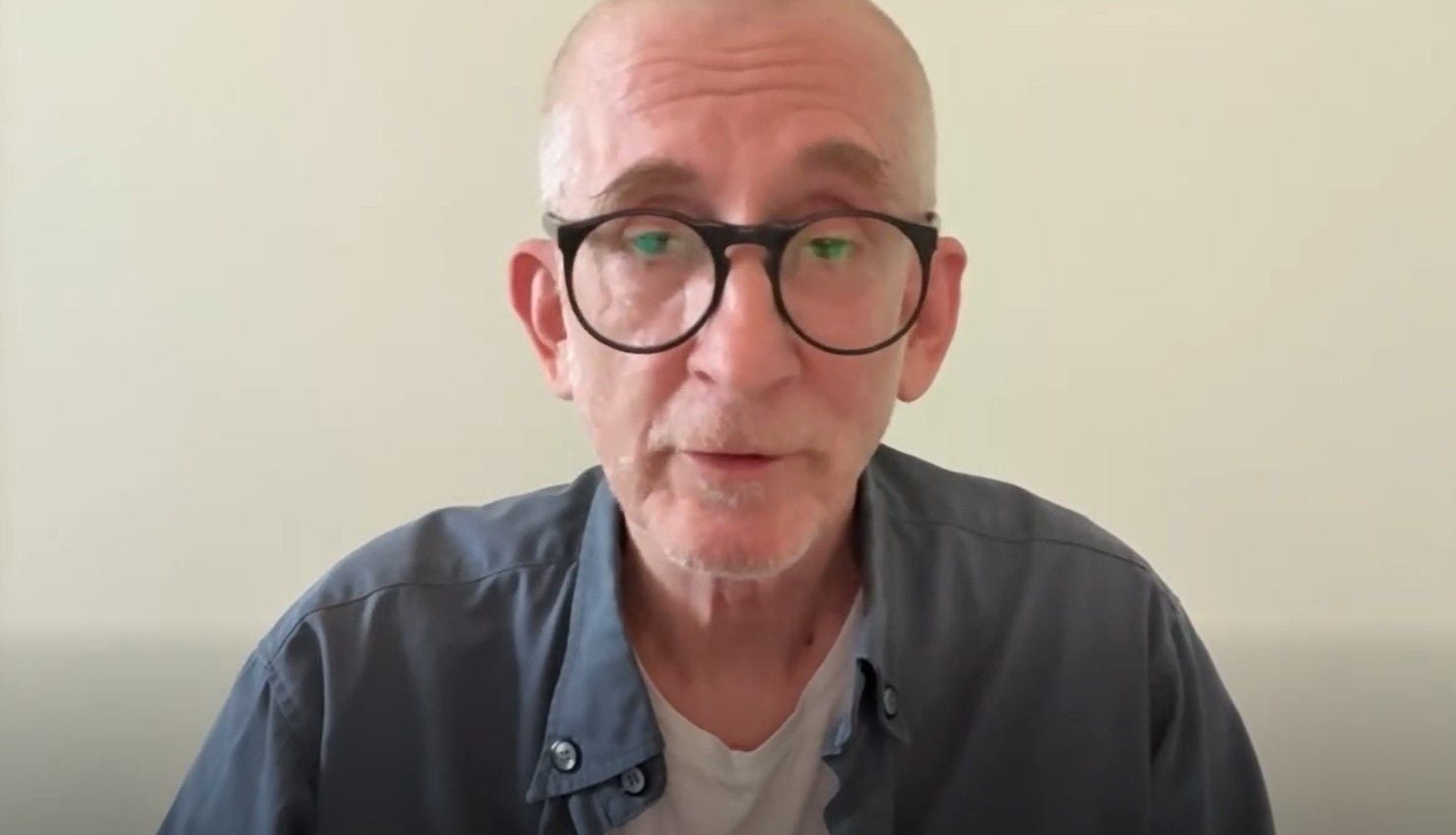 Ein Mann mit Brille und Glatze sitzt vor einer weißen Wand und blickt in die Kamera.