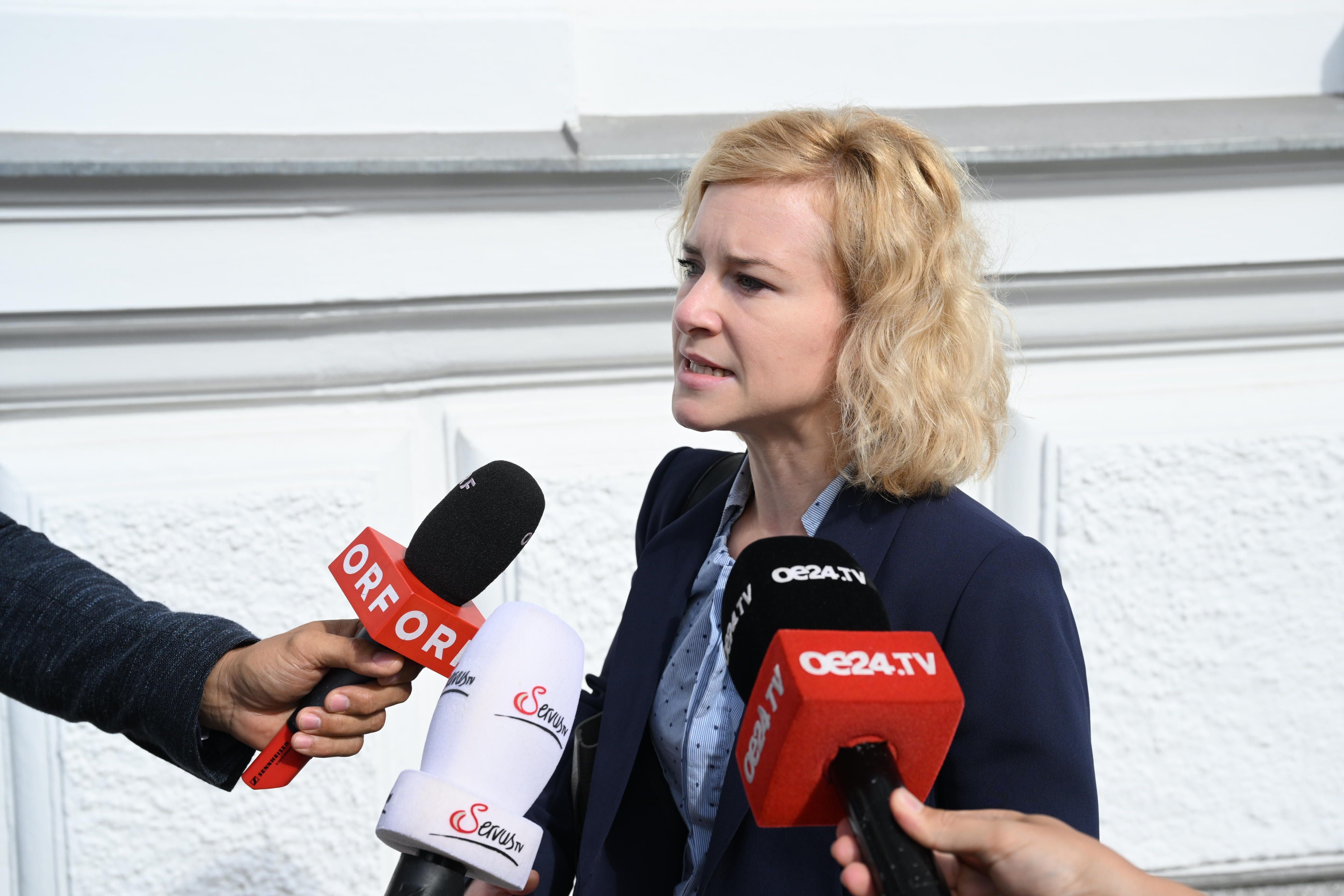 Frau wird vor Mikrofonen von Journalisten interviewt, Hintergrund ist eine weiße Wand.