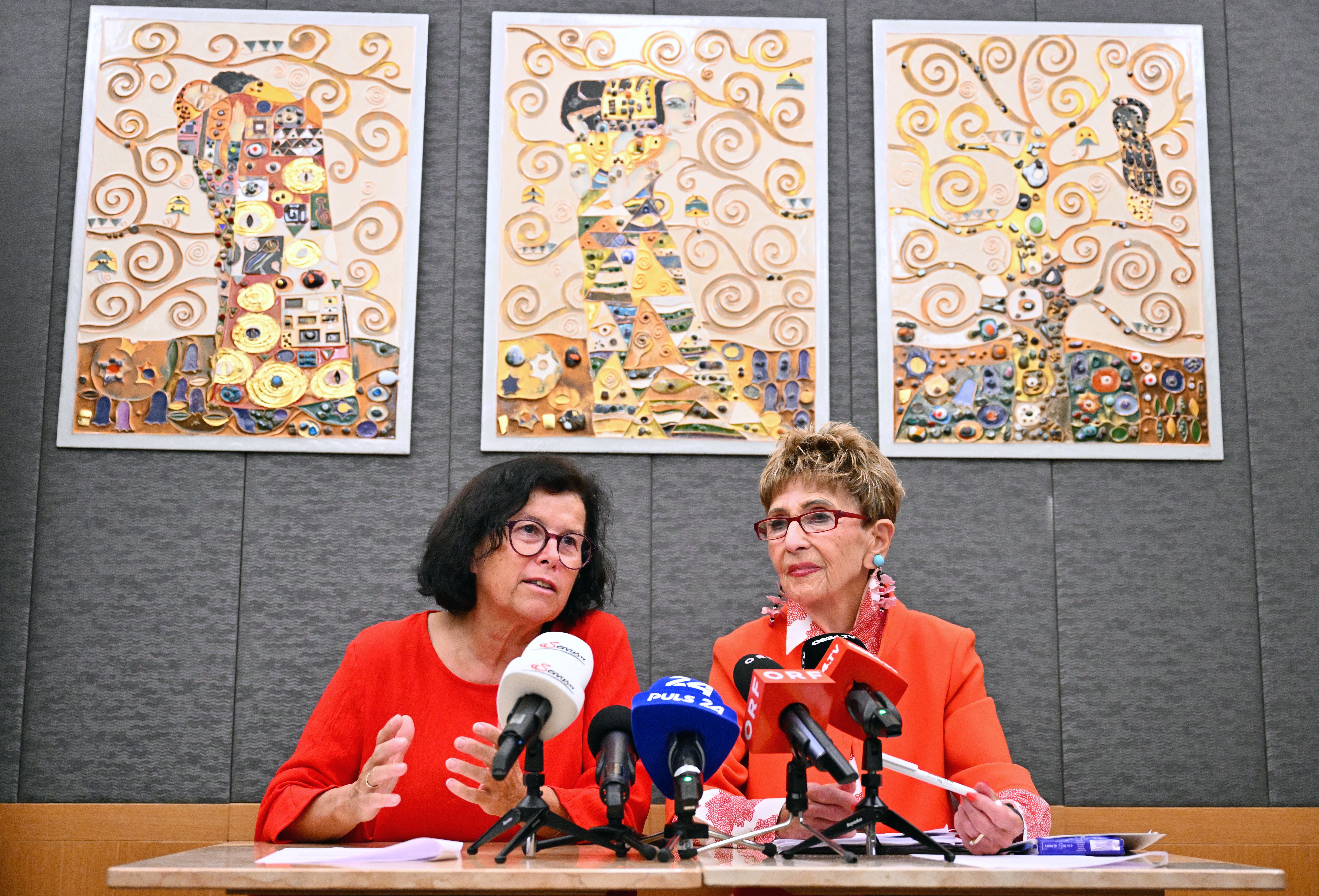 Zwei Frauen bei einer Pressekonferenz mit Mikrofonen, hinter ihnen drei bunte Wandbilder.