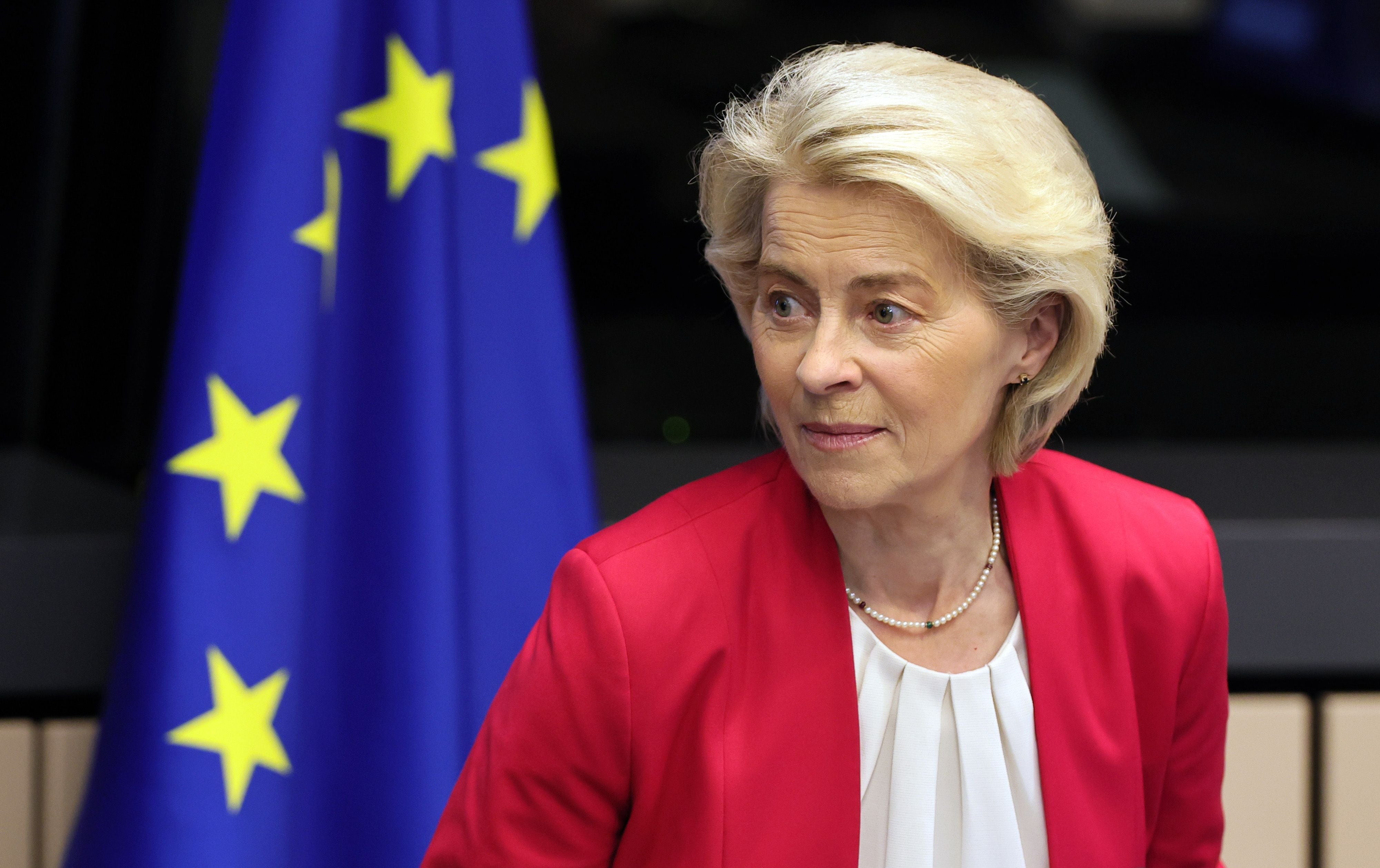 Eine Frau im roten Blazer sitzt vor einer EU-Flagge und blickt zur Seite.