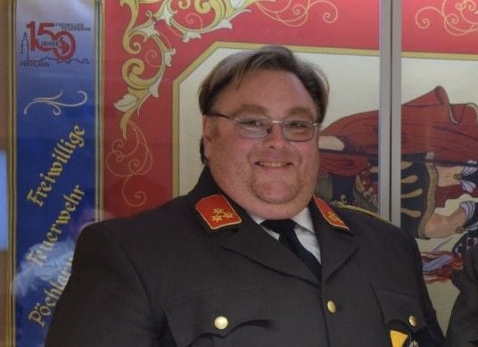 Ein Mann in Feuerwehruniform lächelt in die Kamera, im Hintergrund bunte Banner.