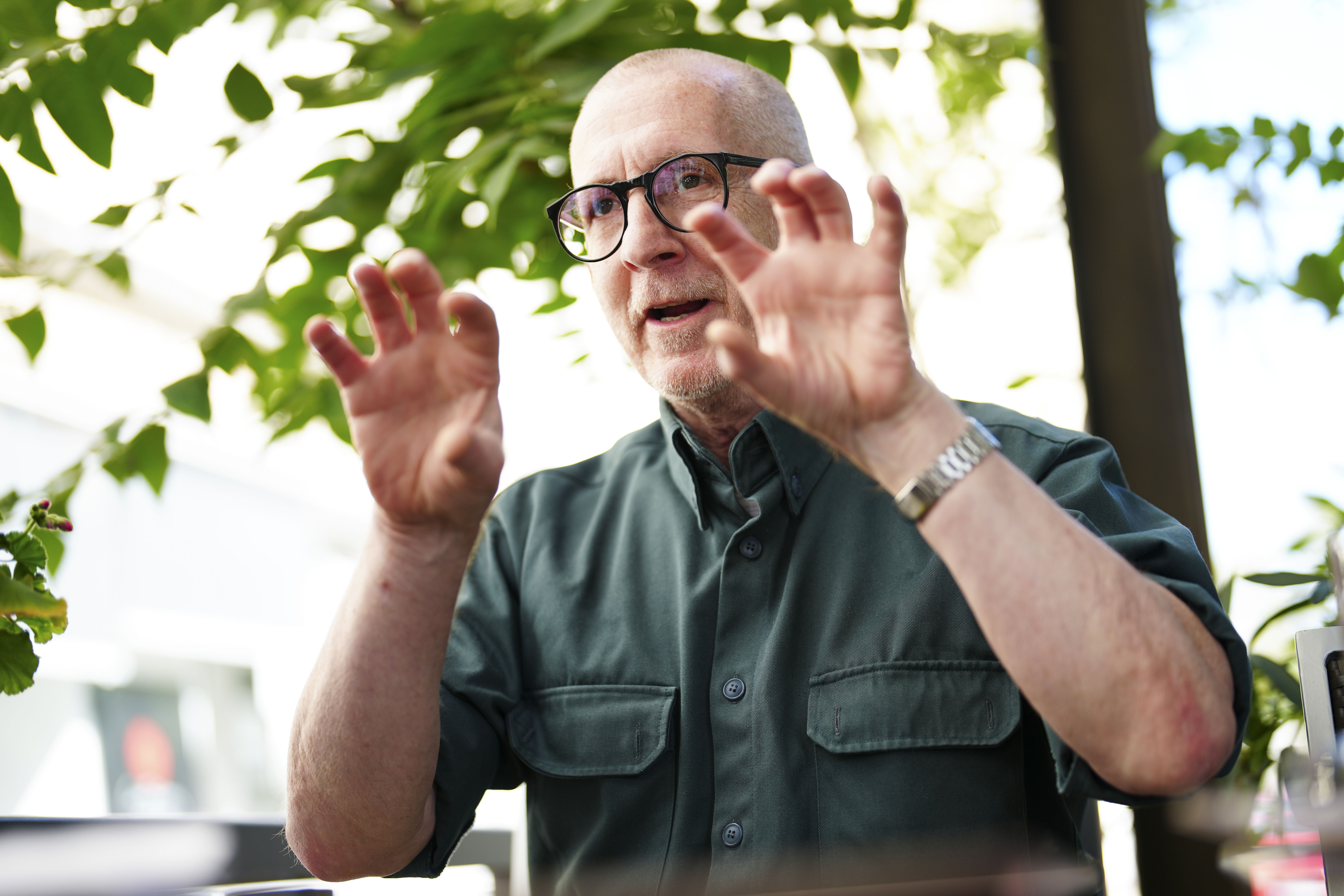 Ein älterer Mann mit Brille spricht gestikulierend im Freien unter grünen Blättern.