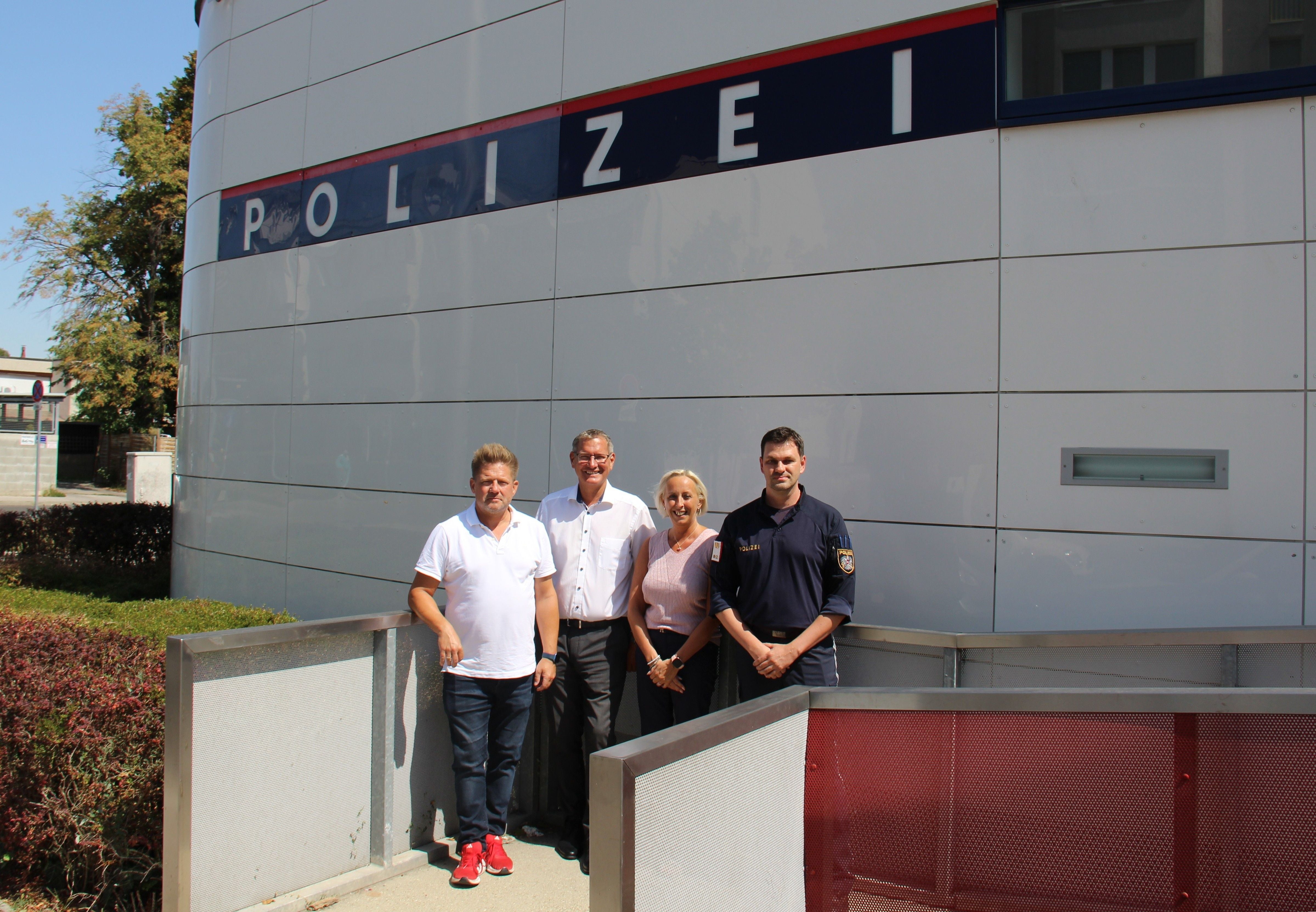 Vier Personen stehen vor einem Gebäude mit dem Schild "POLIZEI" an der Wand.