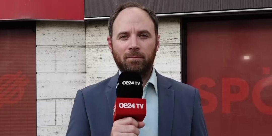Ein Mann im Anzug hält ein OE24 TV Mikrofon und blickt in die Kamera.