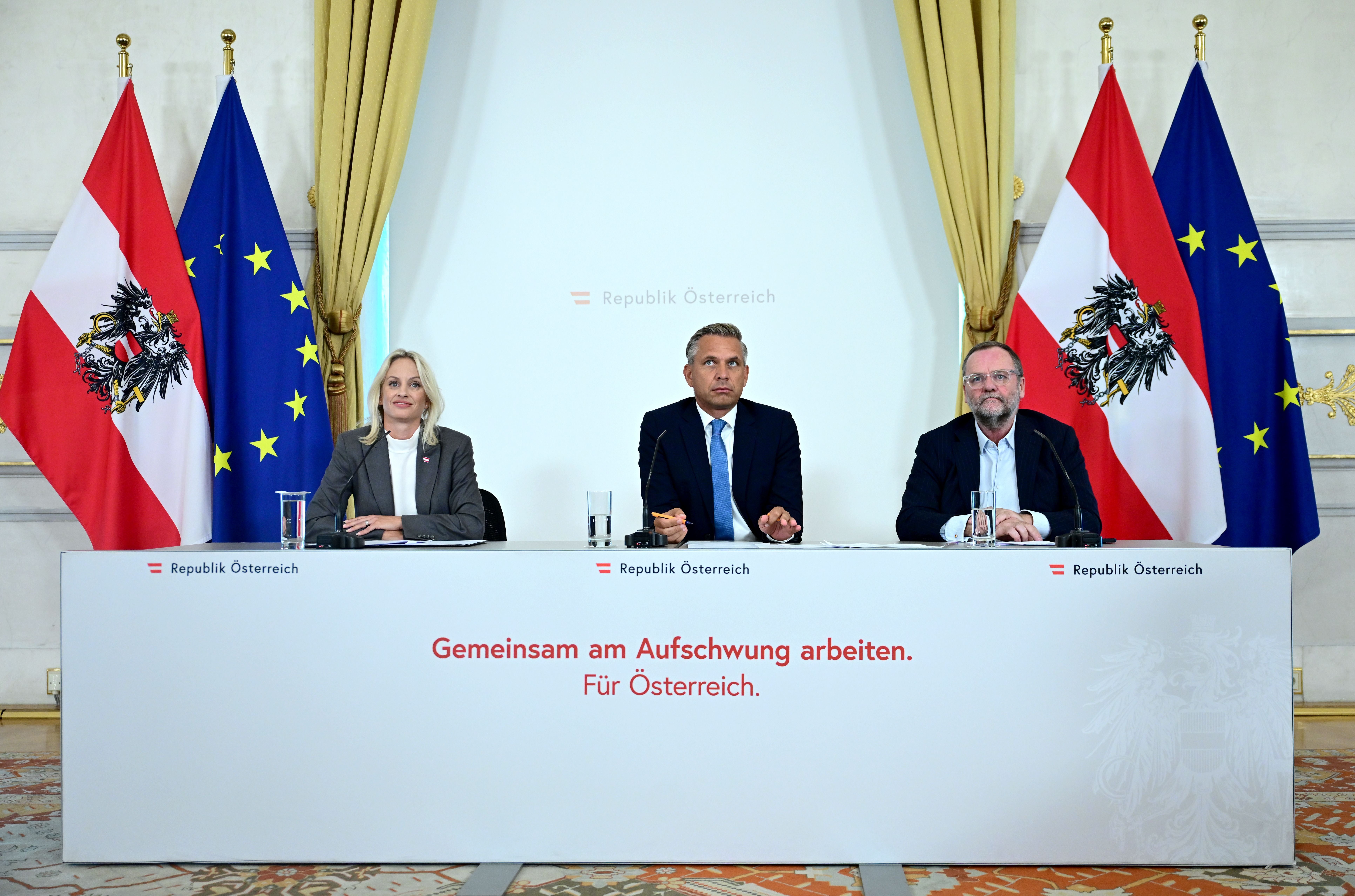 Drei Personen sitzen bei einer Pressekonferenz vor Österreich- und EU-Flaggen.