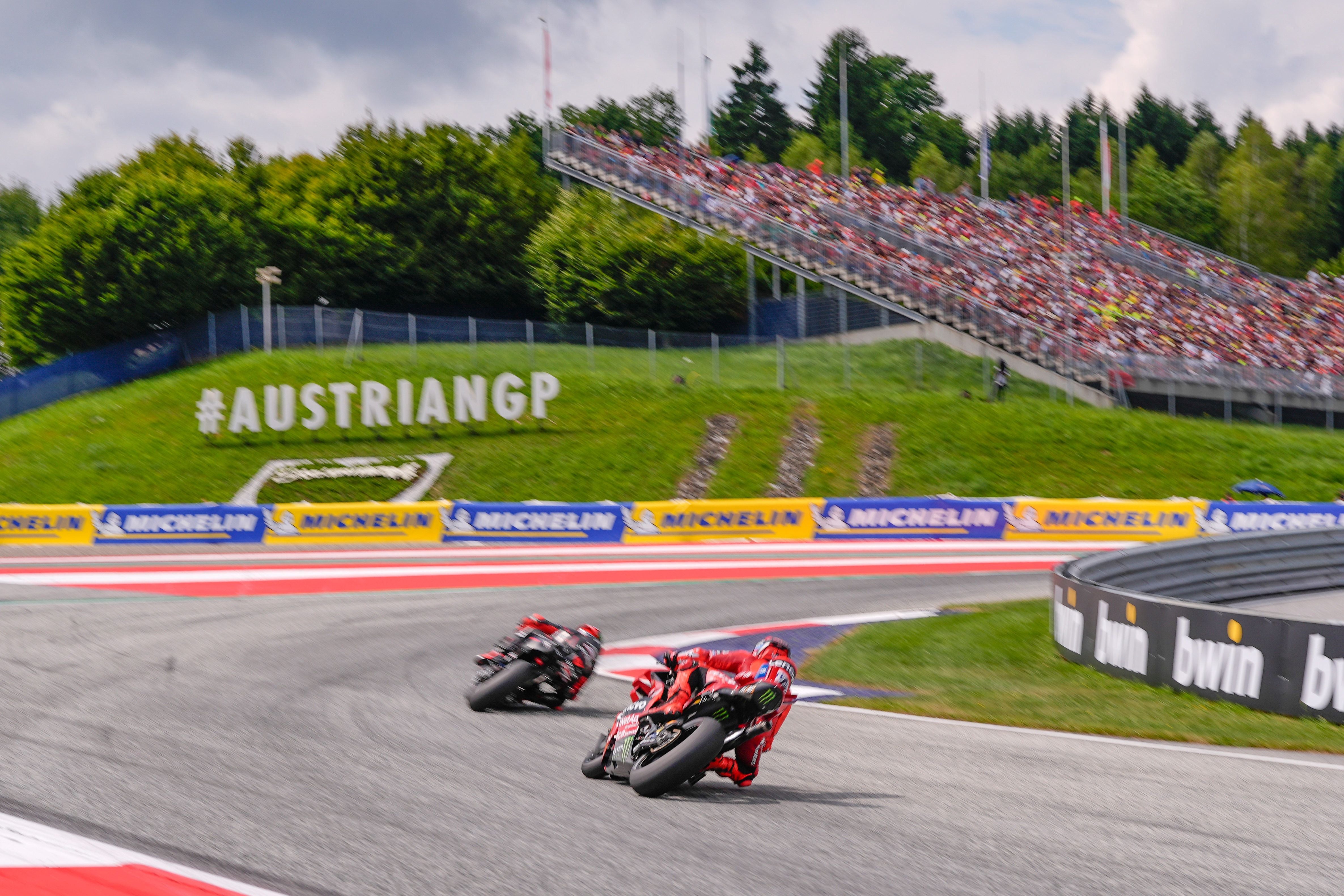 Zwei MotoGP-Fahrer fahren eine Kurve auf dem Red Bull Ring, Zuschauertribüne im Hintergrund.