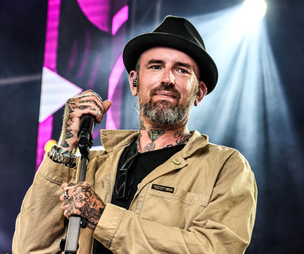 Mann mit Hut, Bart und Tattoos hält Mikrofon auf einer Bühne mit Lichteffekten.