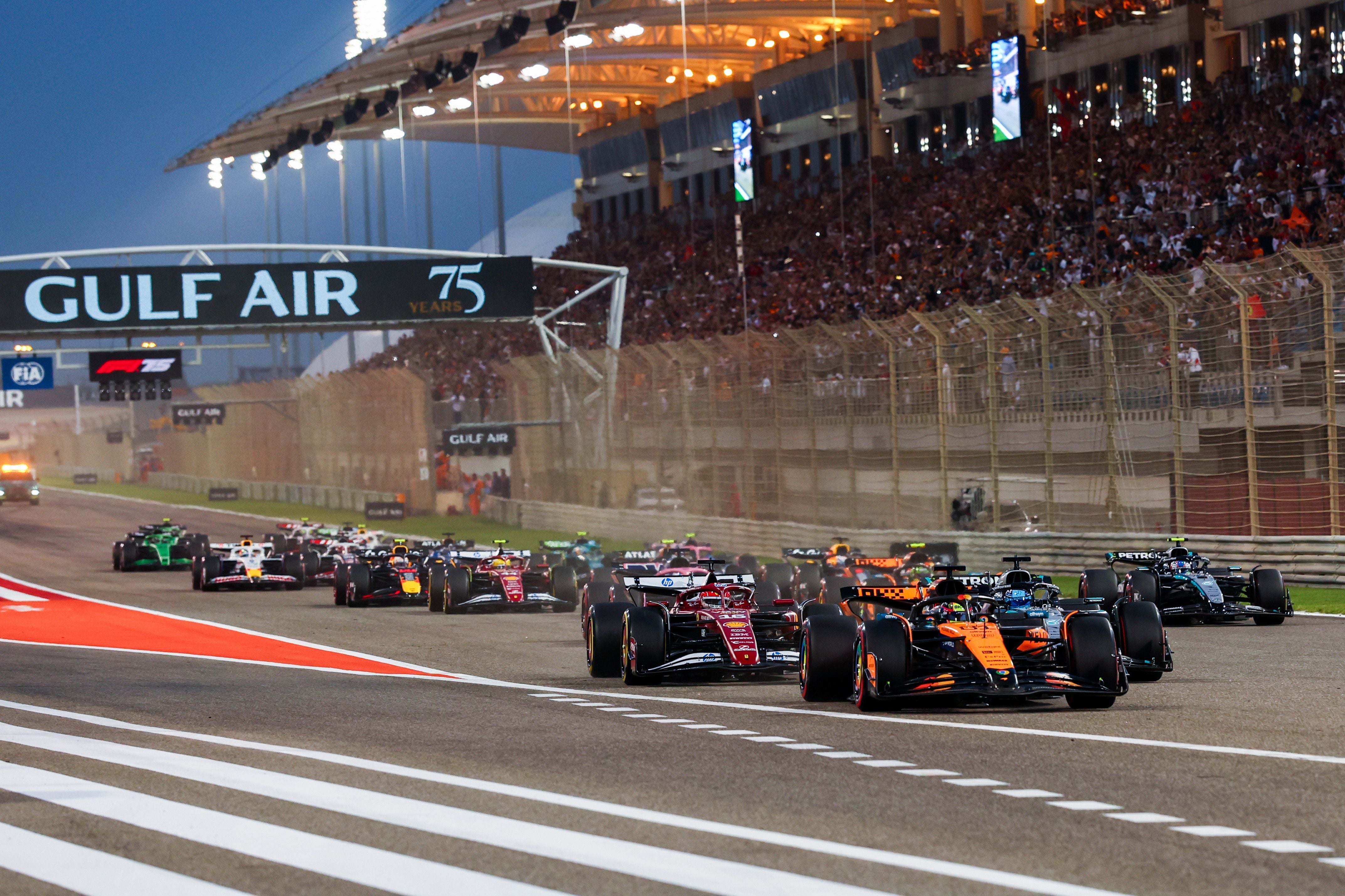 Formel-1-Boliden biegen dicht aneinandergereiht unter dem Flutlicht des Bahrain International Circuits in die Startkurve ein.