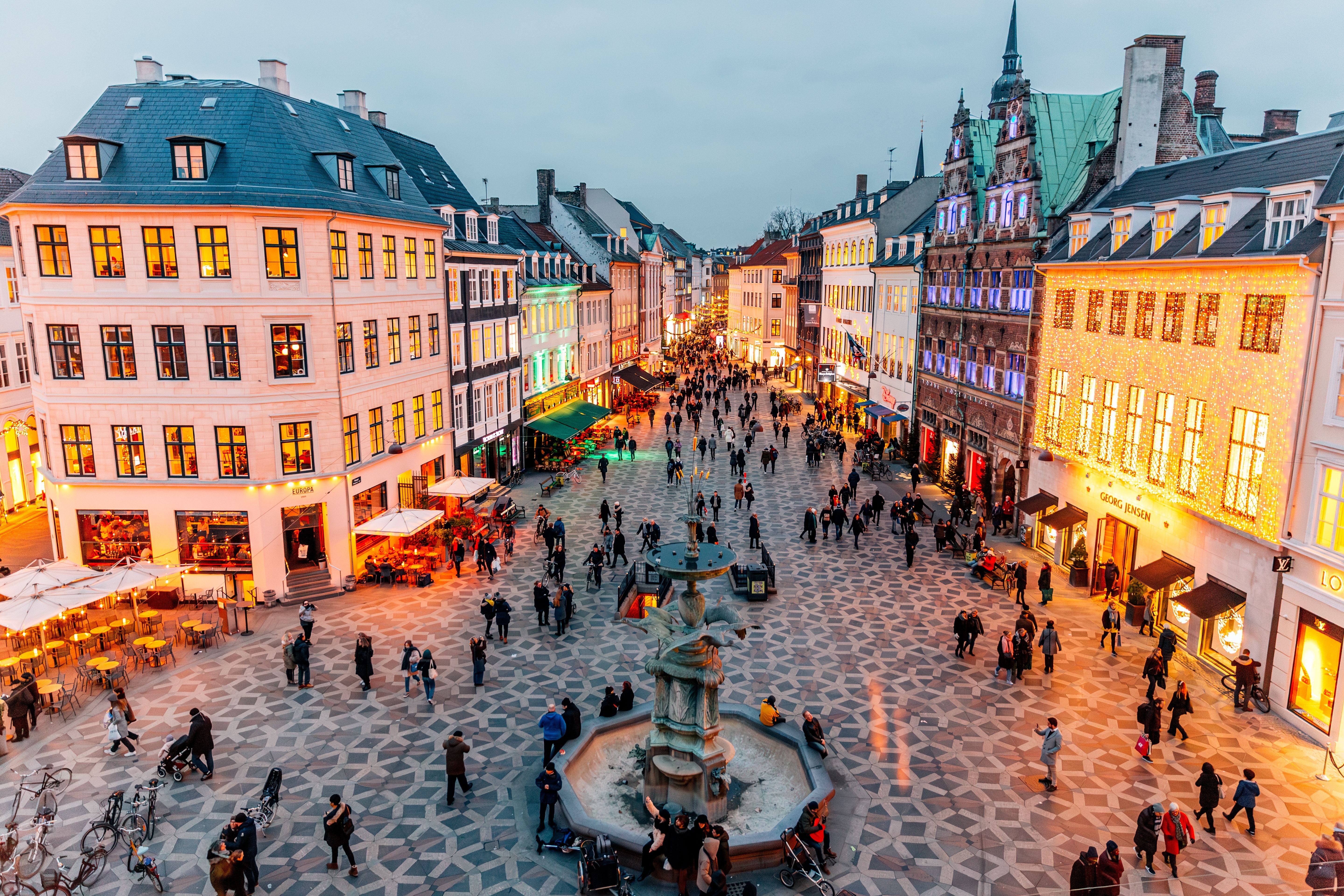 Belebte Fußgängerzone in Kopenhagen mit beleuchteten Gebäuden und Brunnen bei Abenddämmerung.