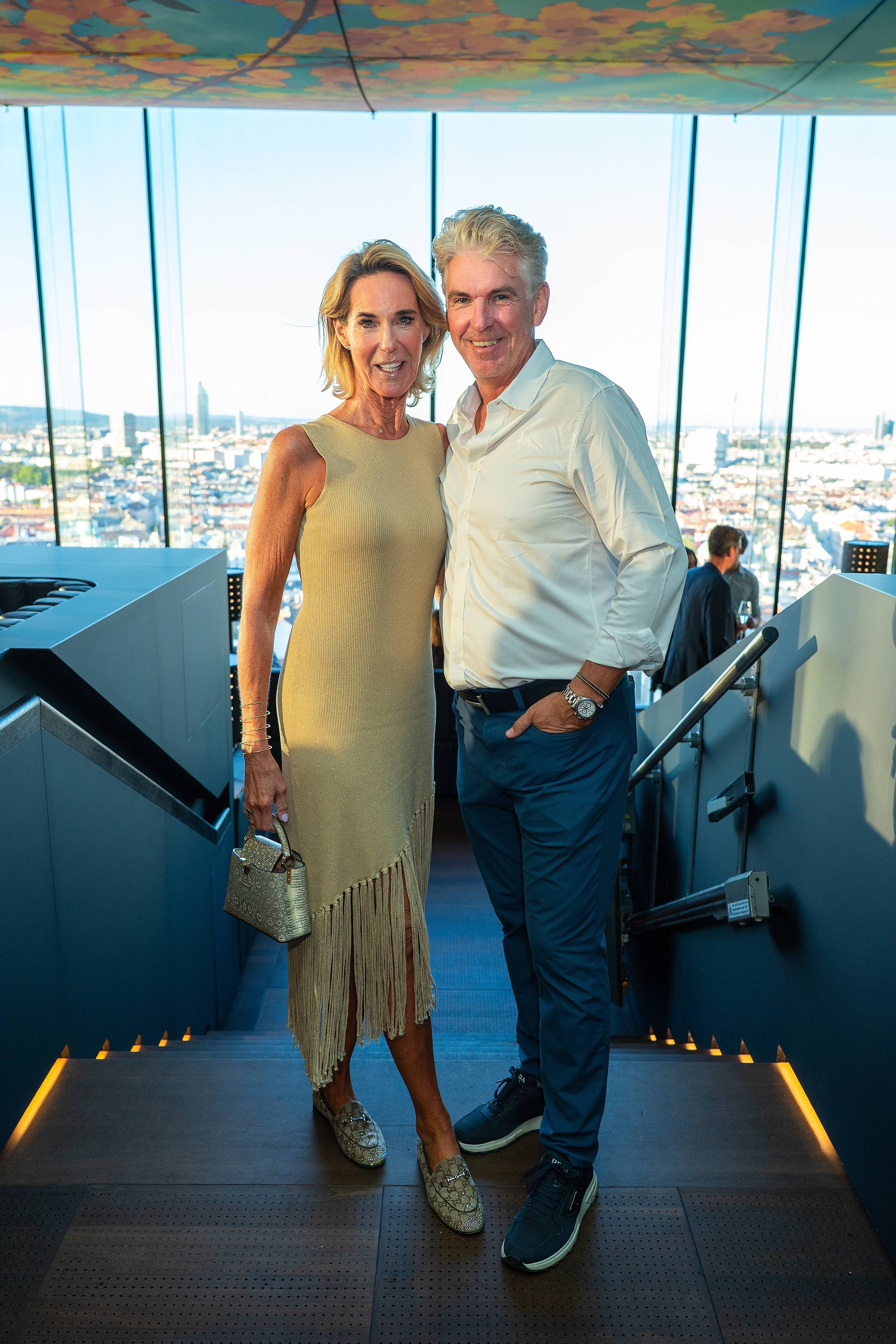 Ein Paar posiert in eleganter Kleidung in einer modernen Rooftop-Bar mit Stadtblick in Wien.