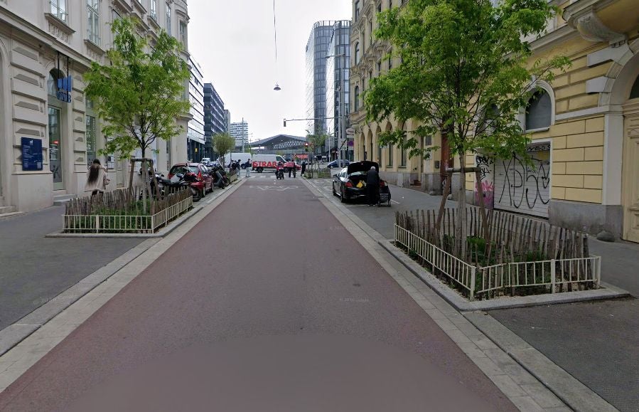 Straße mit Bäumen, parkenden Autos und Menschen, im Hintergrund ein modernes Bahnhofsgebäude.