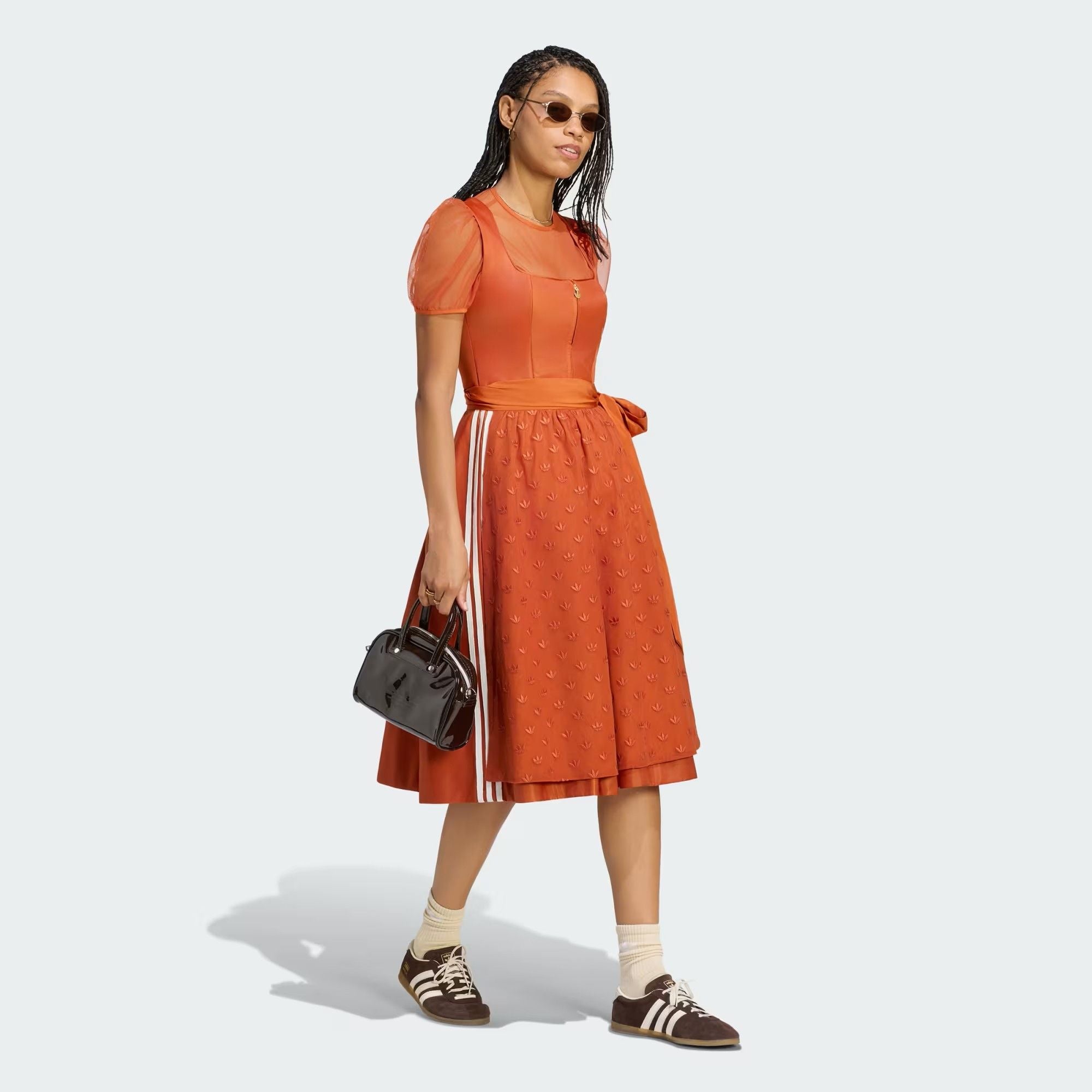 Frau in orangefarbenem Kleid mit Adidas-Schuhen, Sonnenbrille und schwarzer Handtasche.