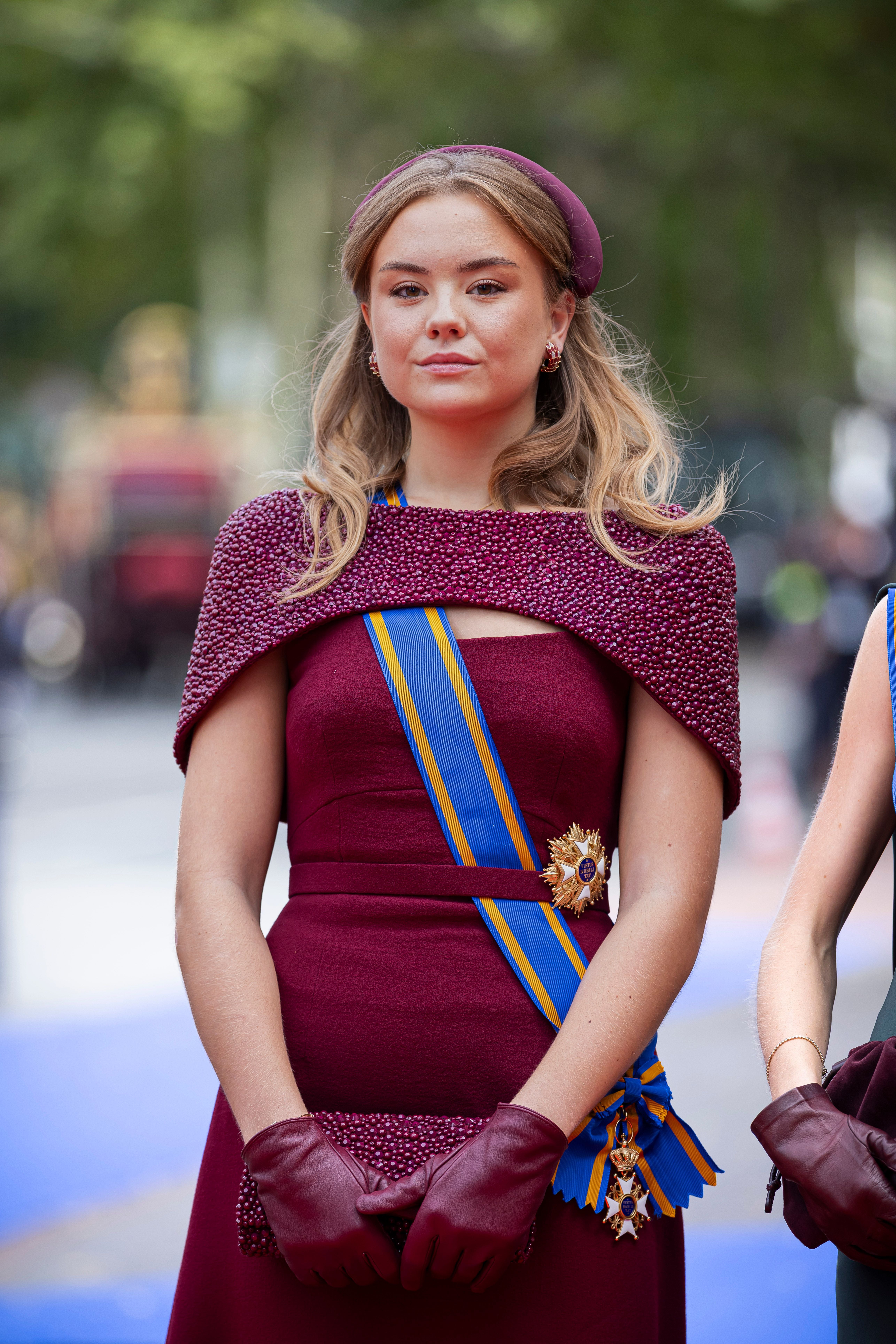 Junge Frau in bordeauxrotem Kleid mit Schärpe und Handschuhen bei einer offiziellen Veranstaltung.