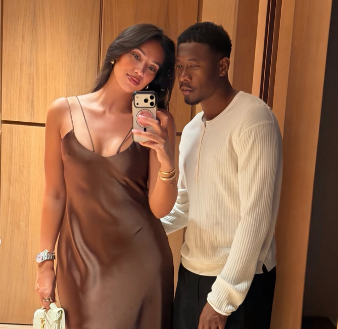 Eine Frau im braunen Kleid macht mit einem Mann ein Spiegelselfie vor Holzpaneelen.