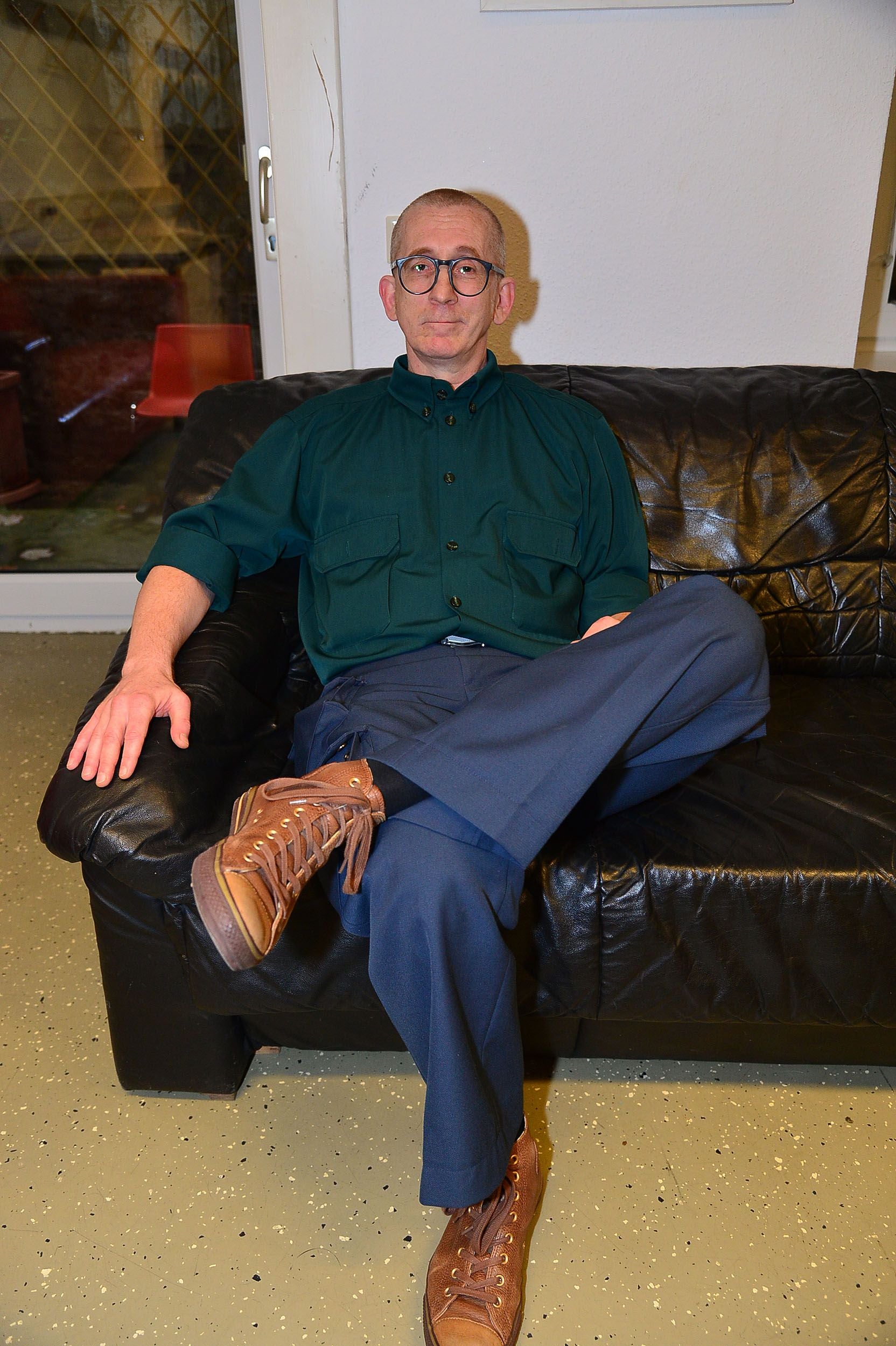 Mann mit Brille sitzt auf schwarzem Ledersofa, trägt grüne Bluse und braune Schuhe.