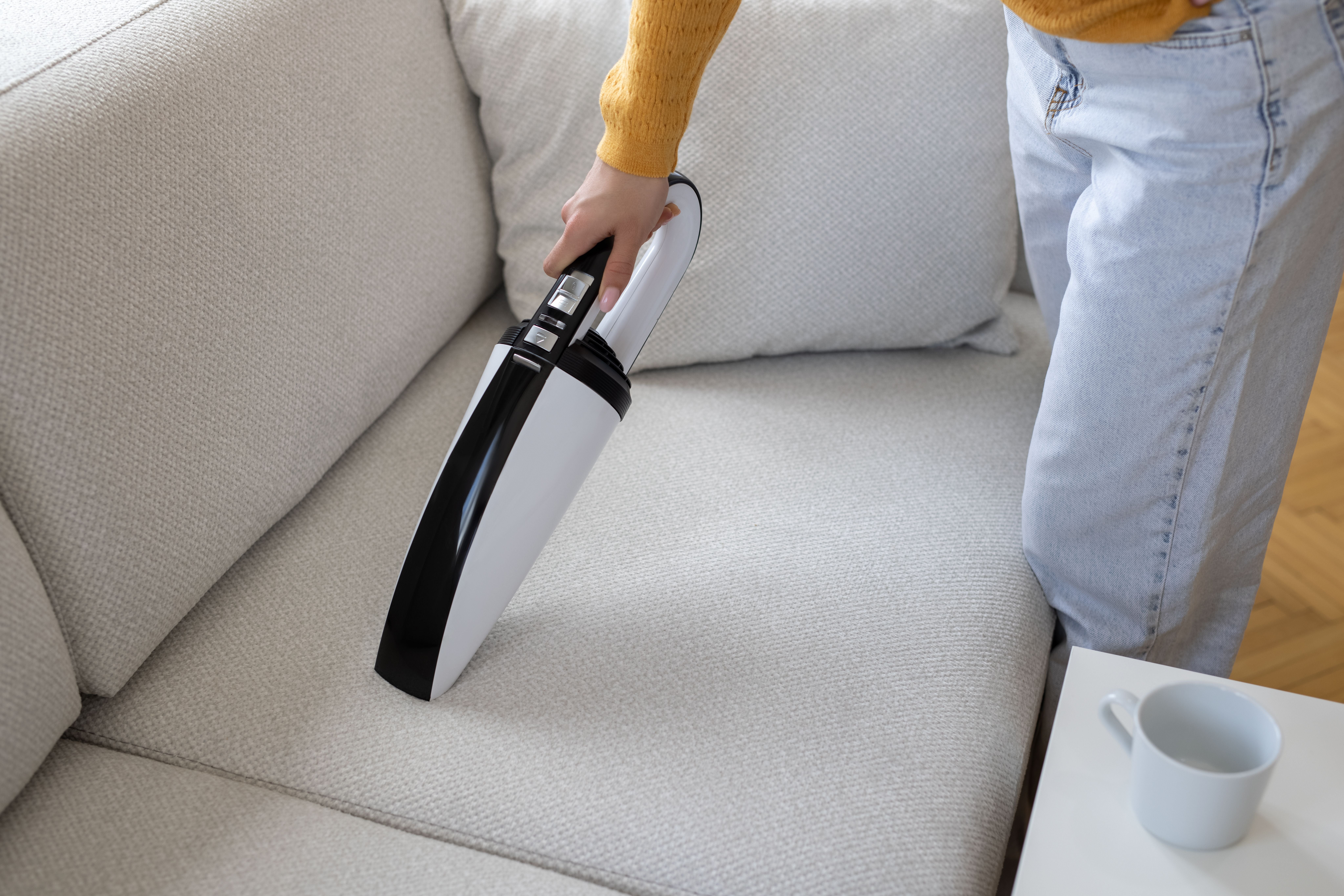 Person saugt mit einem Handstaubsauger ein helles Sofa ab.